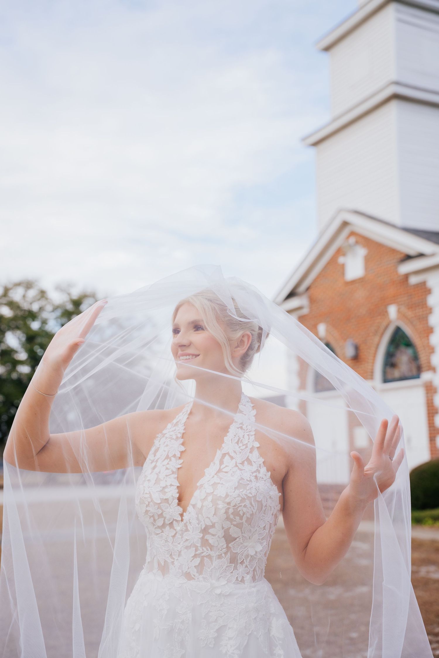 A bride in a delicate lace wedding gown smiles while holding her flowing veil outdoors near a church.
