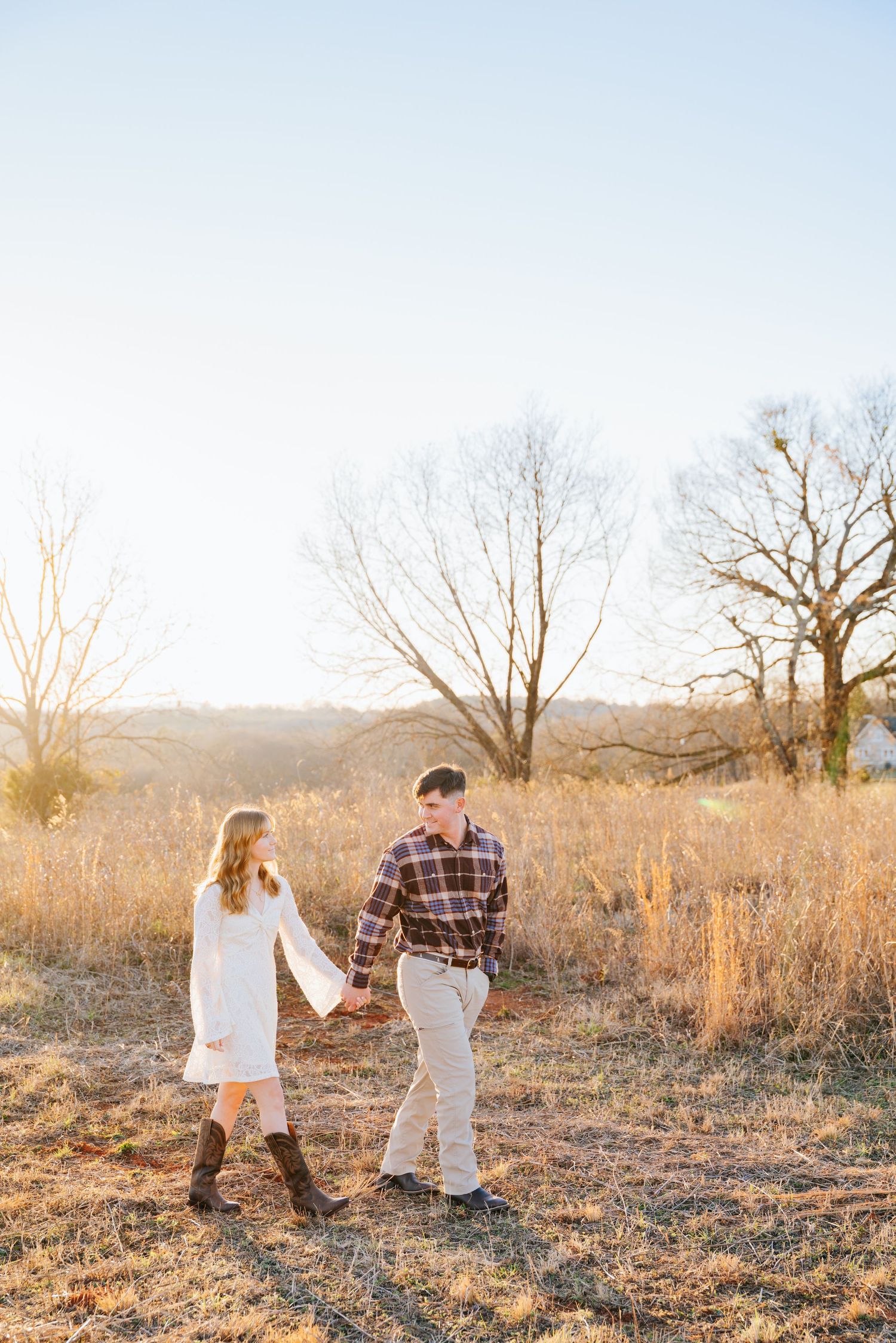 A couple playfully interacts in a golden sunlit field with bare trees in the background during sunset.