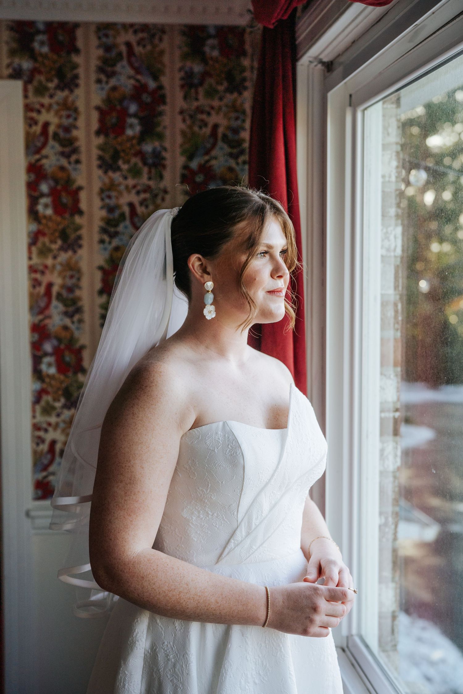 A bride in a strapless gown gazes out a window with floral wallpaper and red curtains.