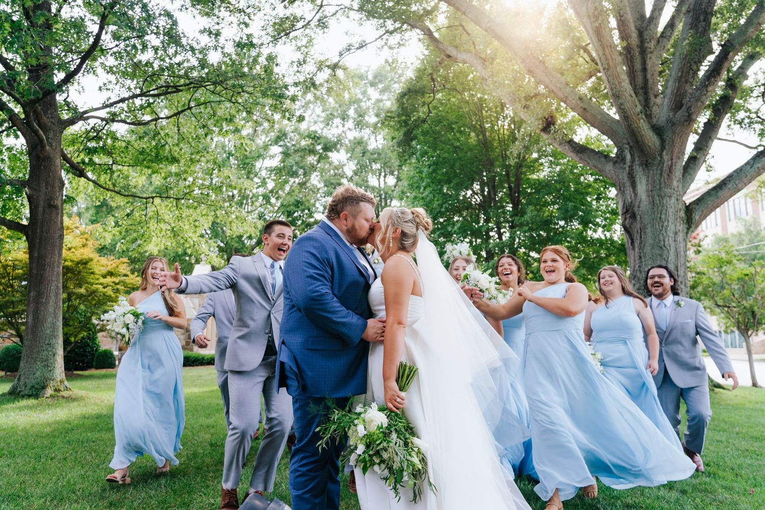 Wedding party in light blue dresses and suits celebrates joyfully on a sunny day outdoors.
