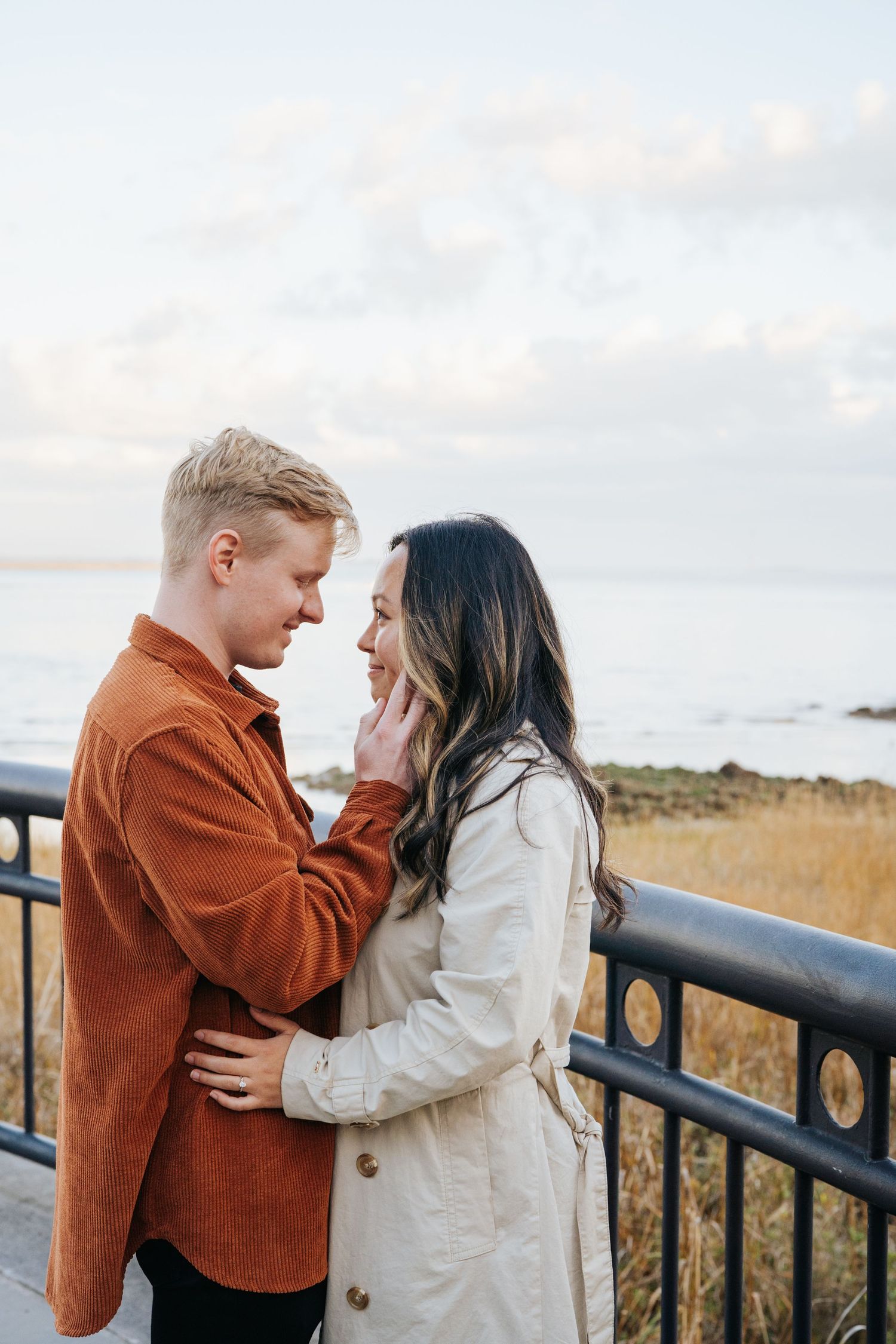 Couple in casual fall attire sharing intimate moment against coastal backdrop.