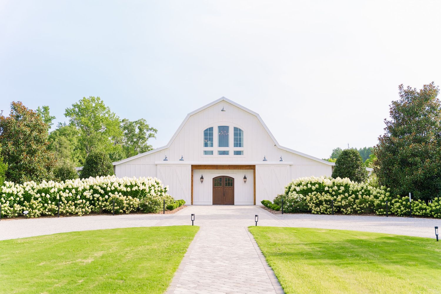 White barn wedding venue with manicured lawn, hydrangea bushes, and a stone pathway leading to wooden doors.