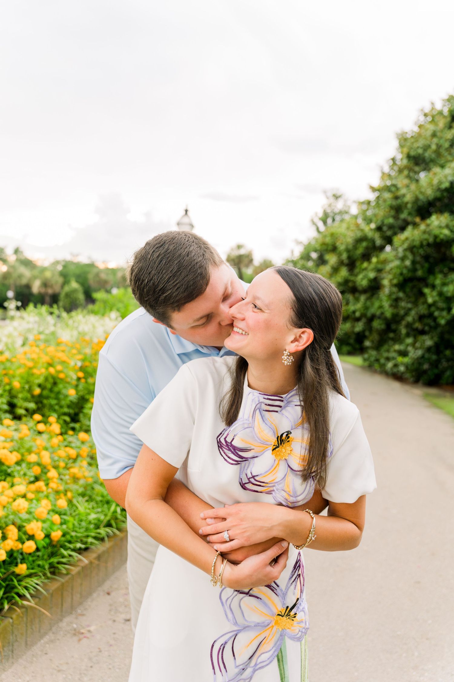 A couple shares a tender kiss outdoors near yellow flowers, the woman smiling in a floral dress while her partner embraces her.