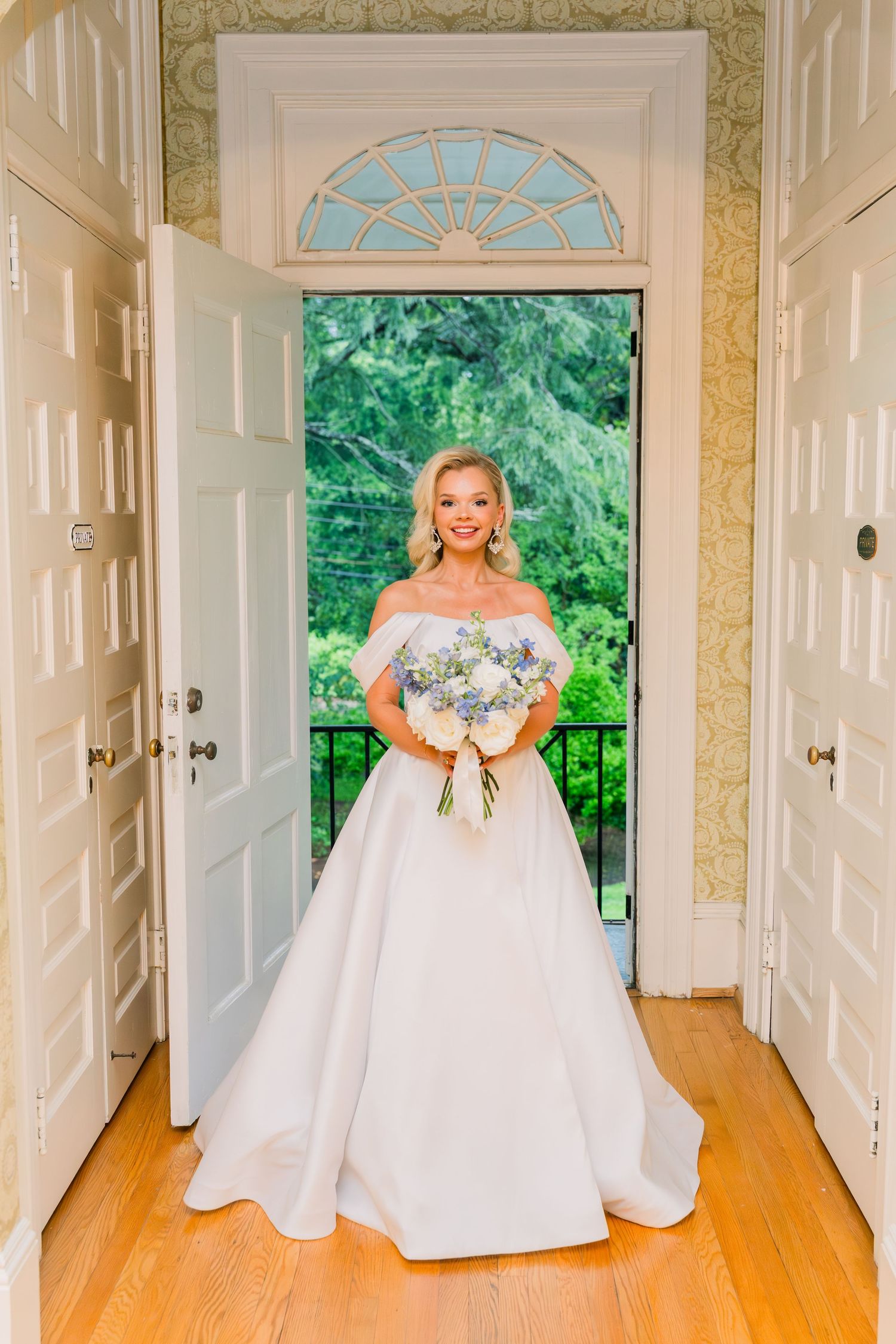 Bride in off-shoulder white ballgown holds blue and white bouquet standing in elegant doorway with garden view.