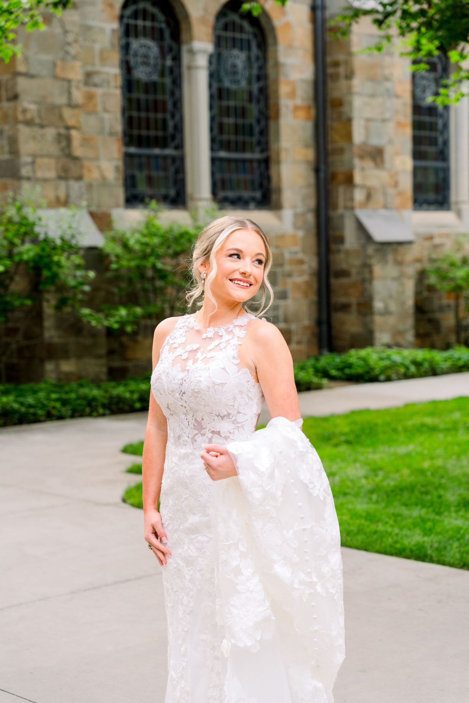 Smiling bride in lace wedding gown holding her dress outside a stone church surrounded by greenery.