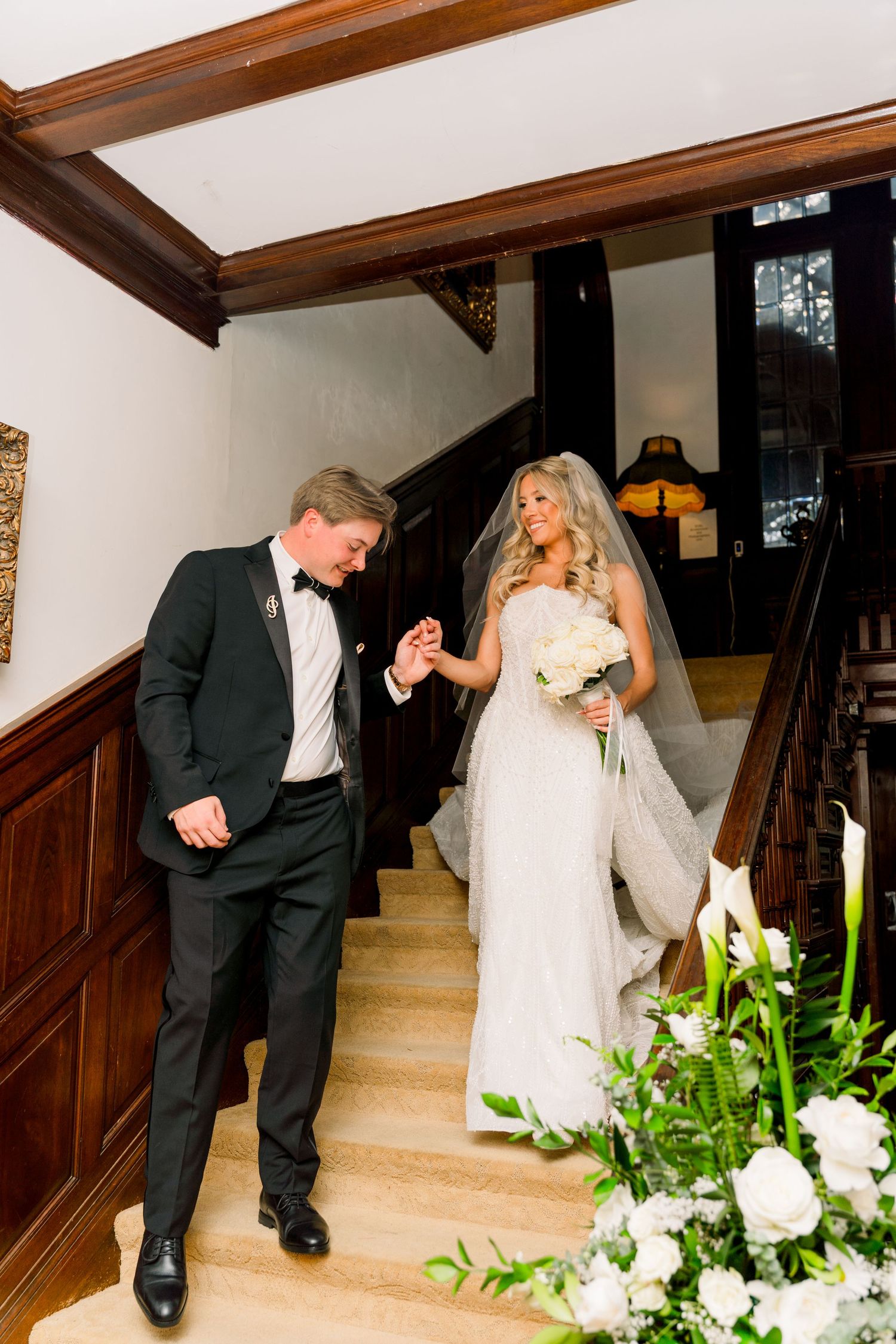Bride and groom descend elegant wooden staircase, bride holding white bouquet in wedding gown with veil, groom in black tuxedo.