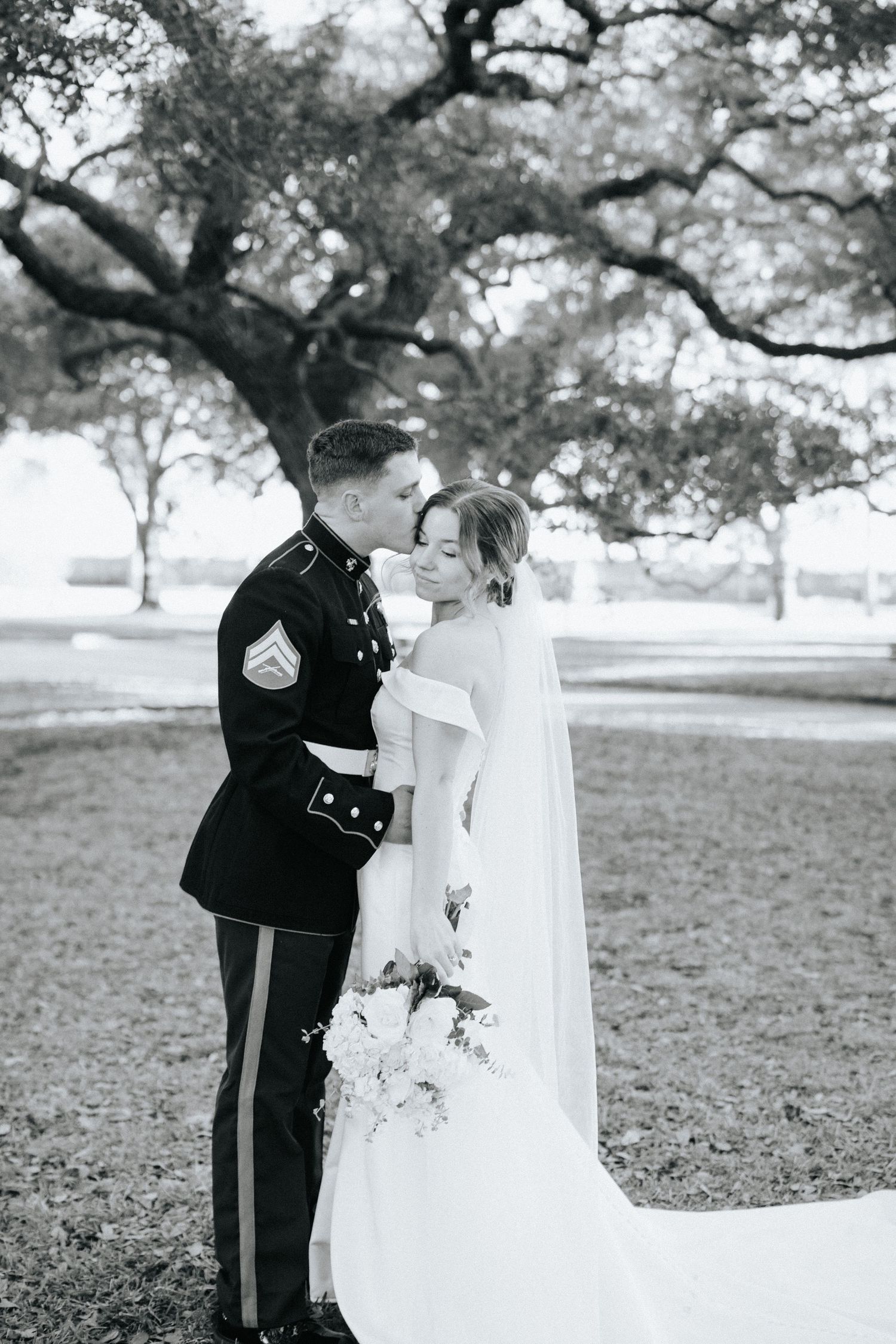 Black and white photo of a couple in formal wedding attire under oak trees.