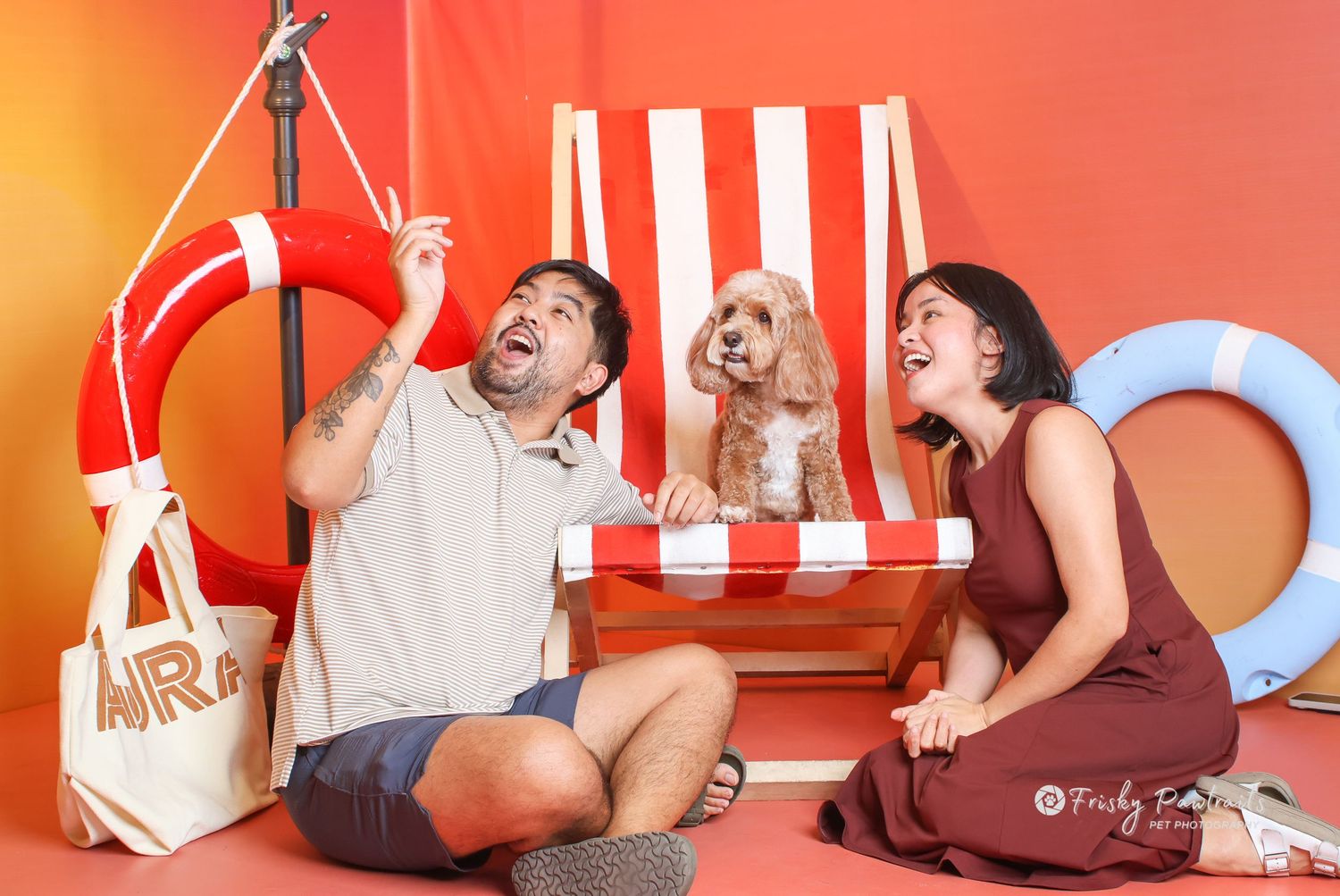 Laughing couple with a fluffy dog posing in a colorful beach-themed photo shoot with striped chair.