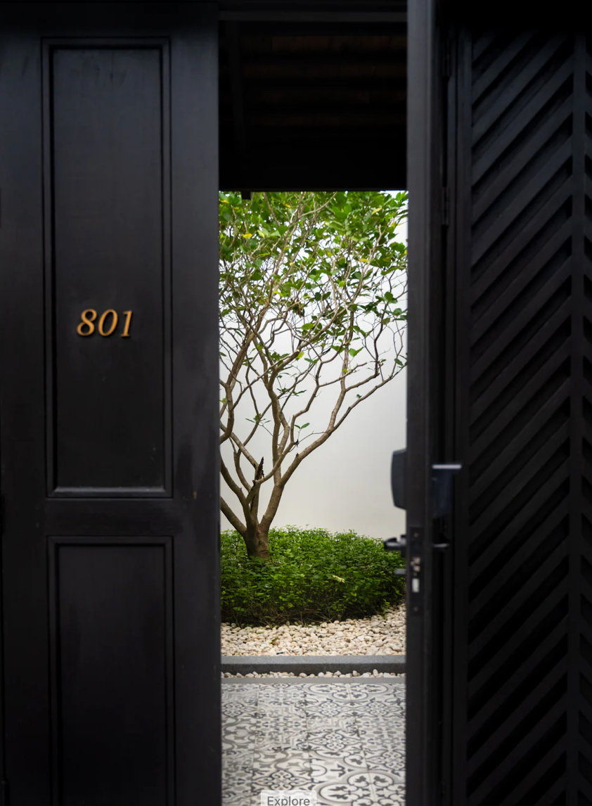 Modern black doors with gold room numbers in minimalist hotel hallway design leading to an interior garden.