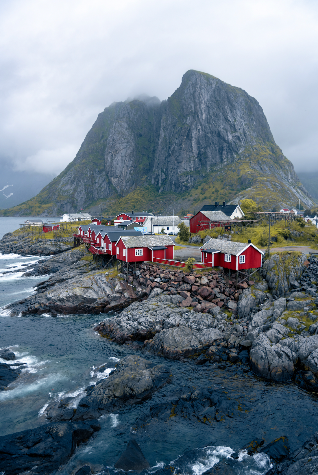Red fishing cabins perched on rocky shoreline with dramatic mountain peaks in Lofoten Islands, Norway.