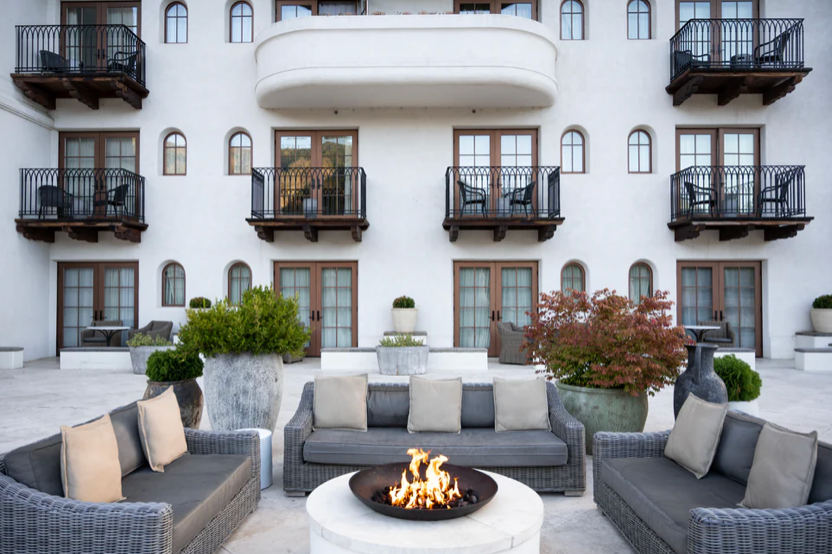 Modern outdoor hotel lounge area with gray furniture and fire pit against white building facade.