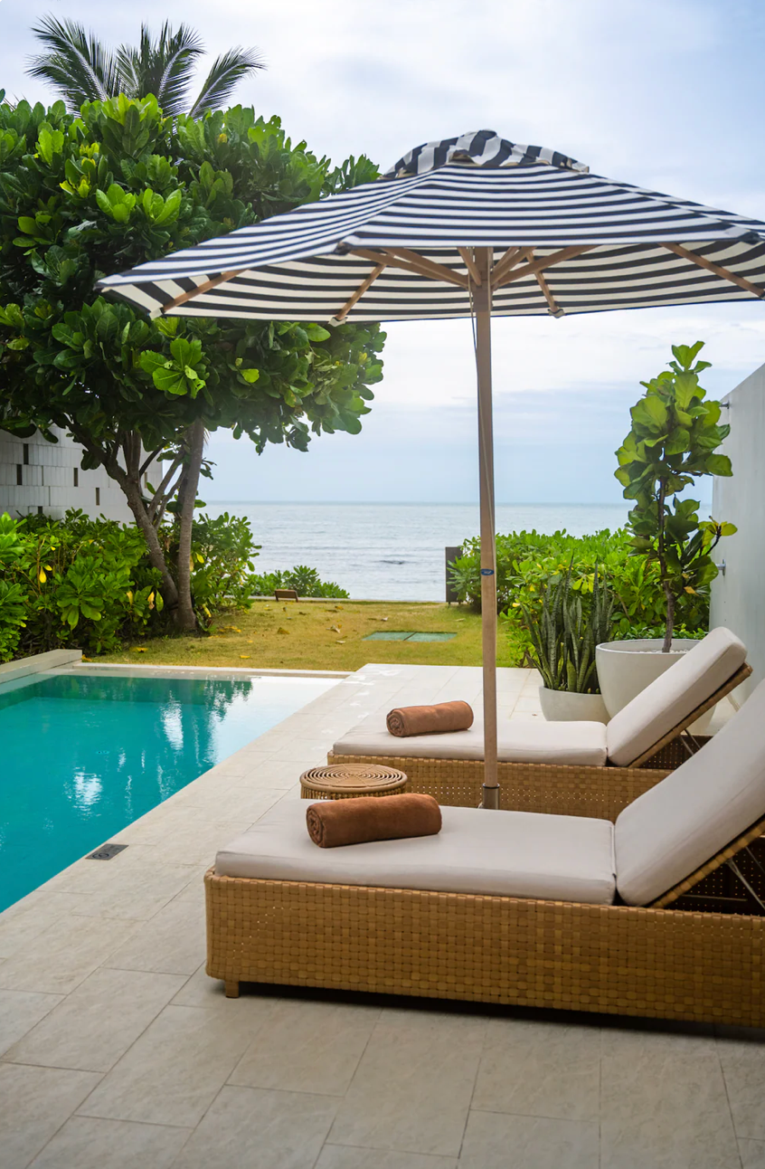 Luxurious poolside loungers with striped umbrellas overlooking tropical oceanfront at Kimpton Kitalay hotel in Koh Samui, Thailand.