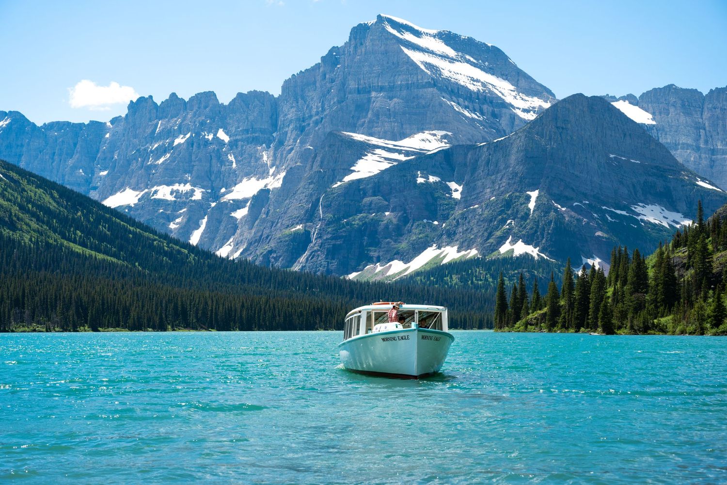 A small boat cruises on turquoise alpine lake waters in Glacier National Park surrounded by snow-capped mountain peaks in Montana.