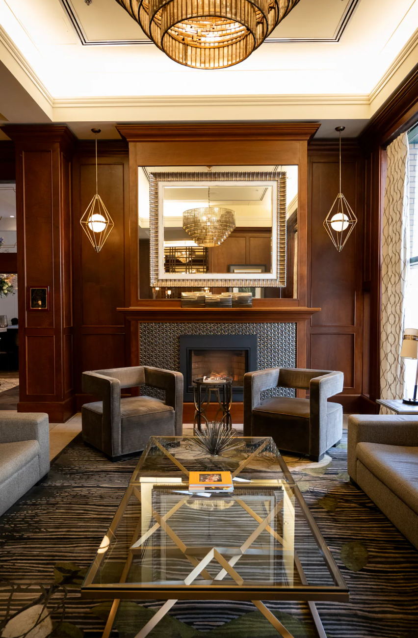 Elegant hotel lobby lounge with wood paneling, modern chandelier, gray armchairs and glass coffee table.