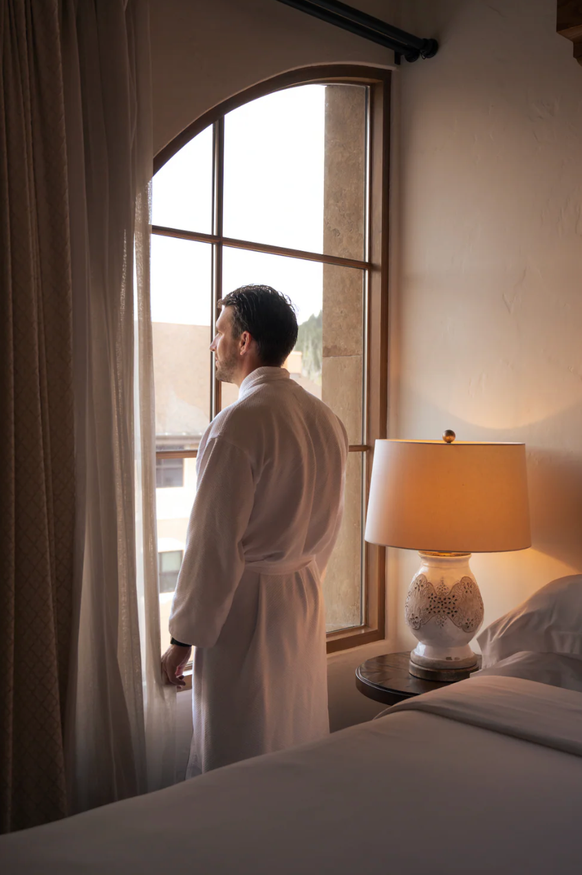 Silhouetted male figure in white robe standing by window in sunlit hotel room.
