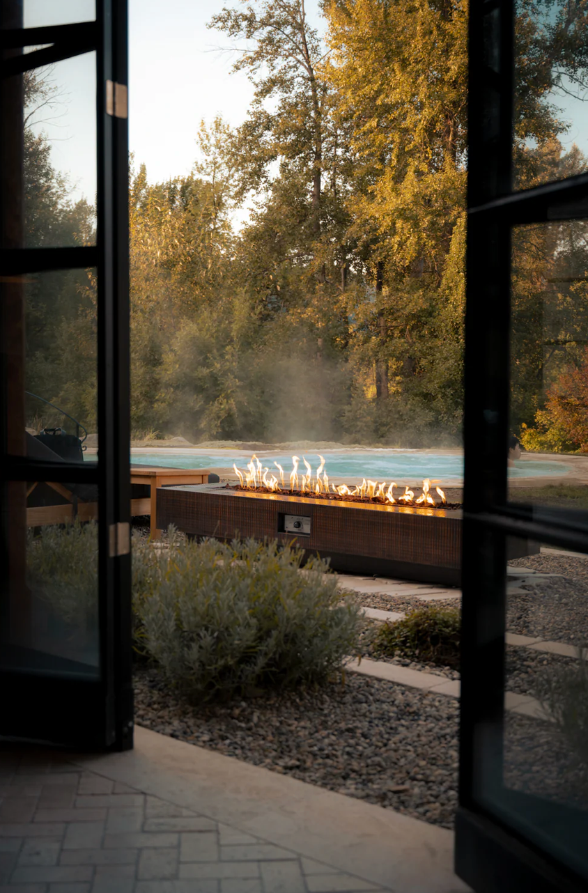 View through glass doors of outdoor fire feature at sunset with Fall trees in background.