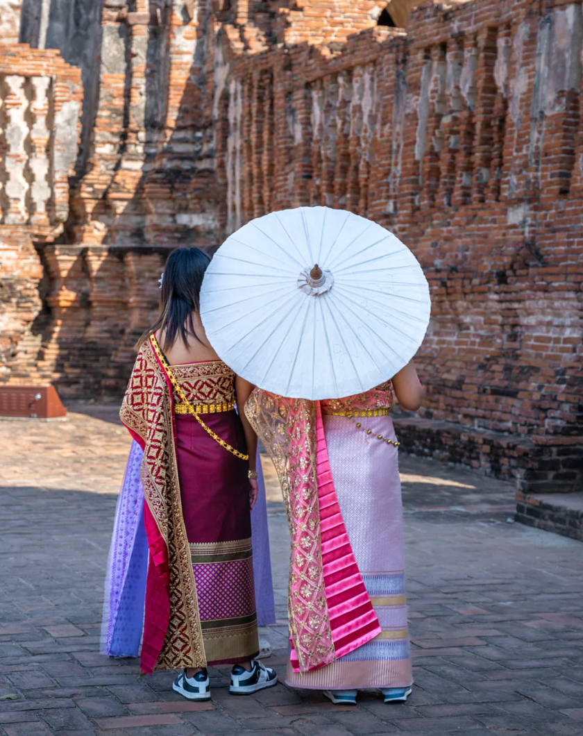 Traditional Thai parasols creating shade for two women against ancient brick temple walls in Thailand.