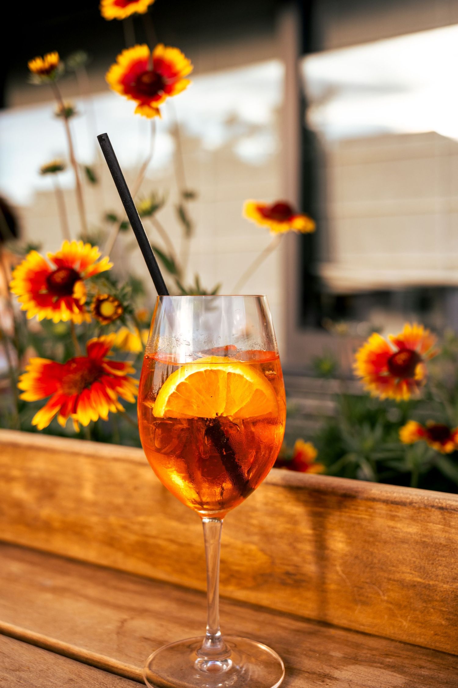 A refreshing Aperol Spritz cocktail with orange slice garnish sits on wooden table with vibrant gaillardia flowers in background.