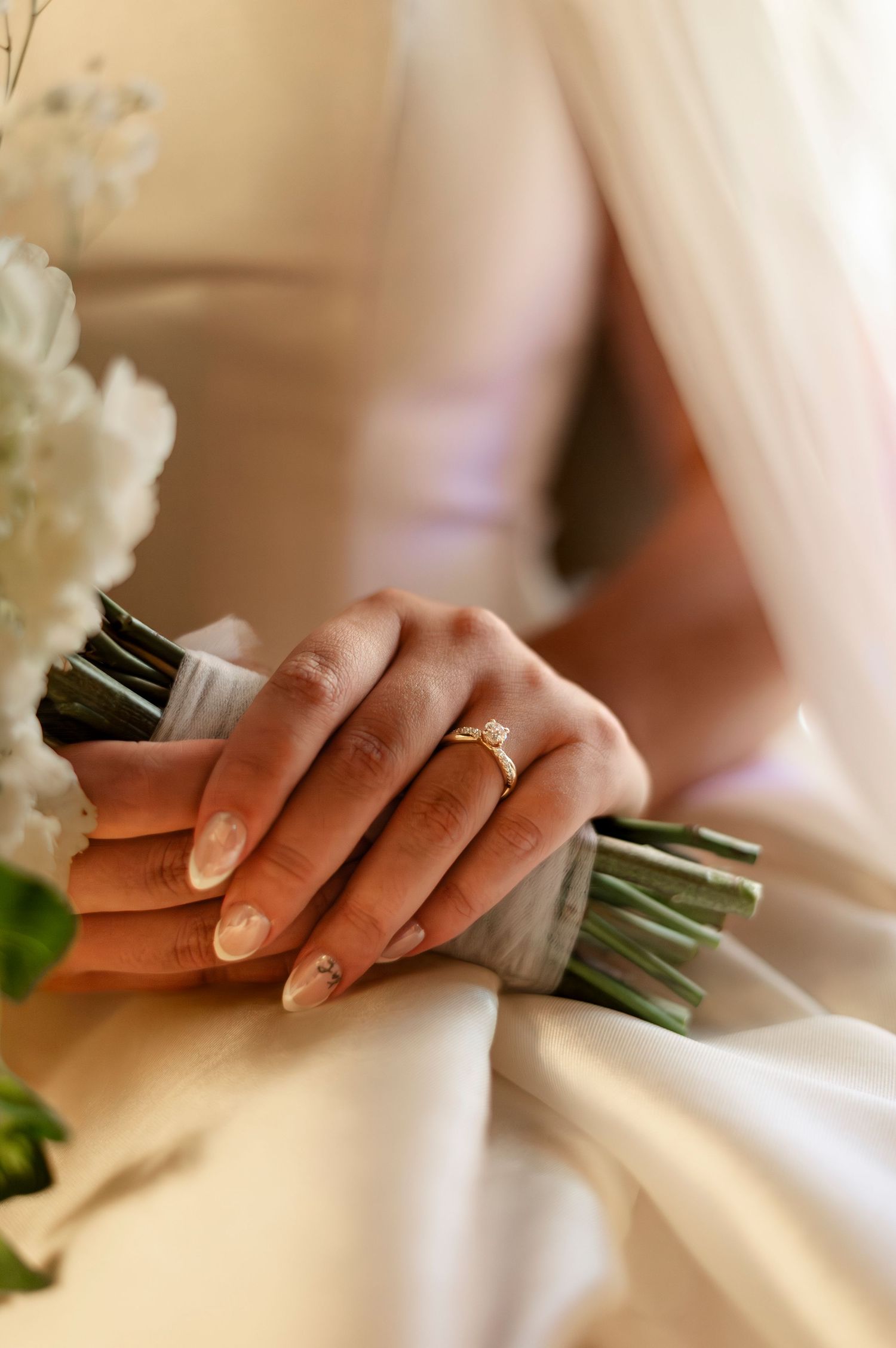 Bride holding white floral bouquet with gold diamond engagement ring on her finger, wearing an elegant white wedding dress.