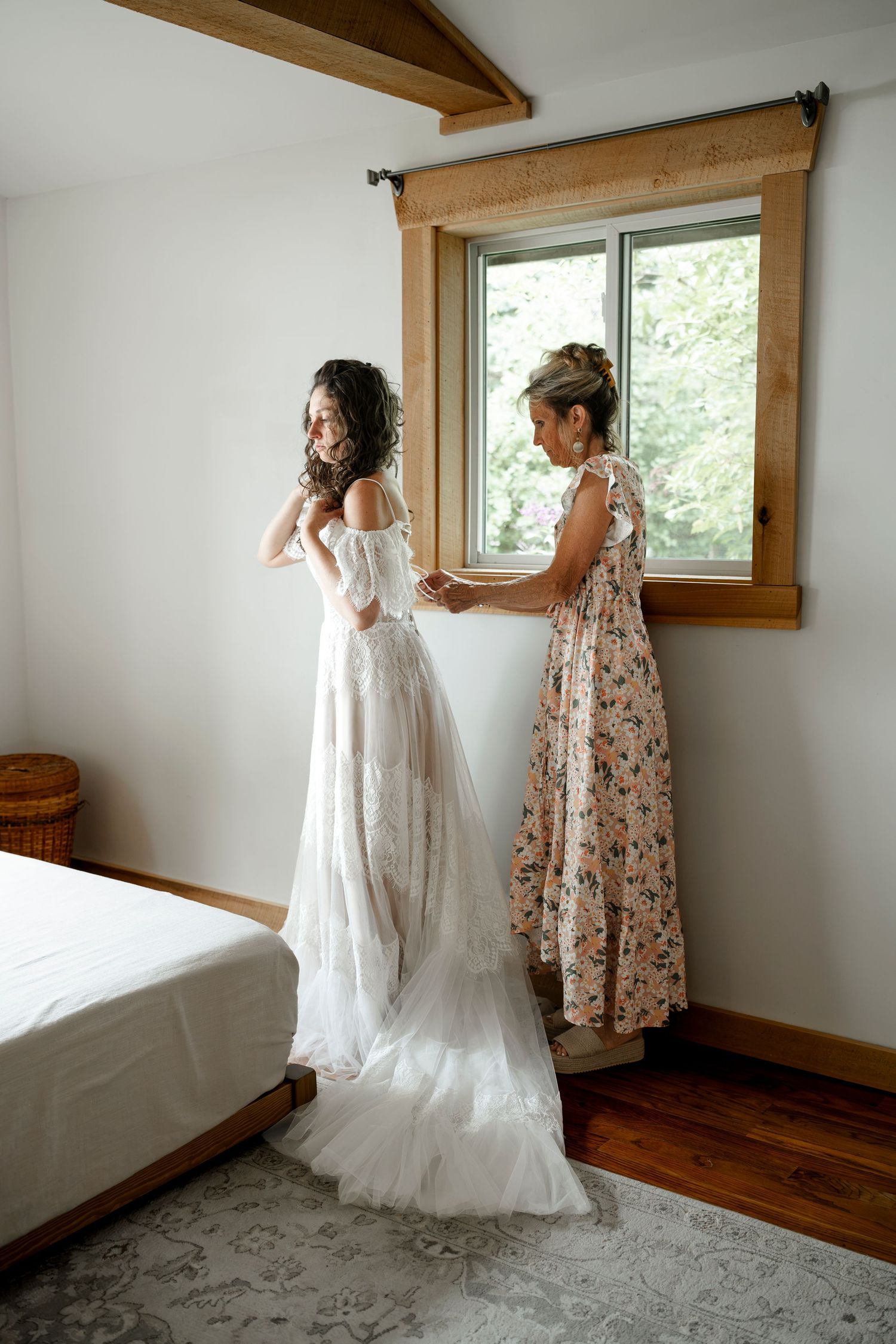 Bride in lace wedding gown gets helped by bridesmaid in floral dress in a bright bedroom.