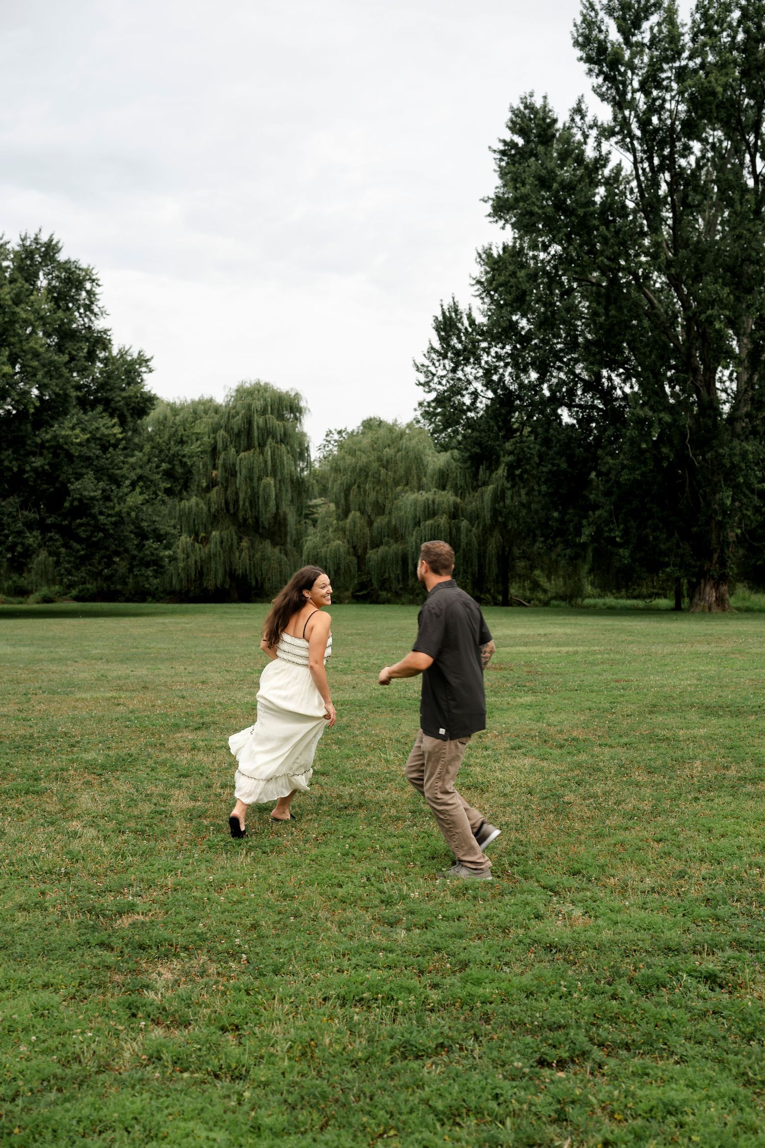 Couple running and holding hands in a lush green park with willow trees in the background.