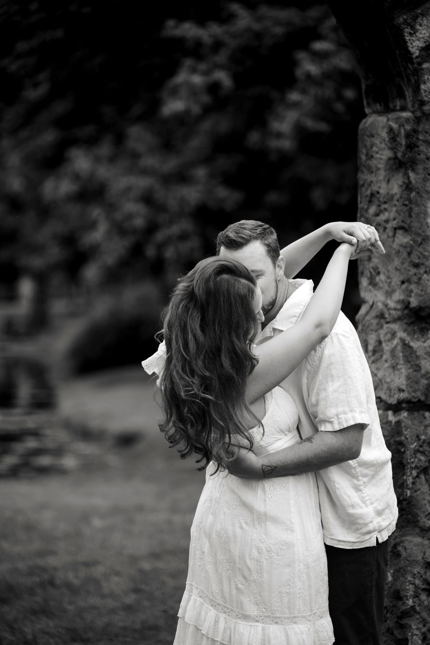 Black and white photo of a couple embracing and kissing near a stone pillar in a lush garden setting.