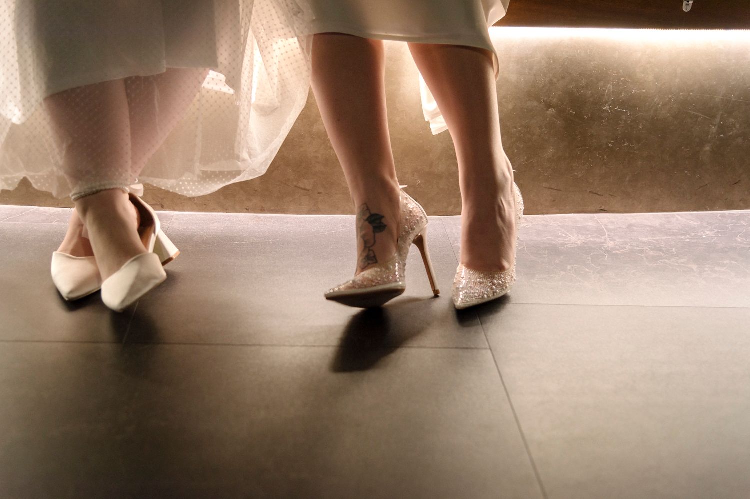 A series of elegant white shoes and dress hems photographed on a dark floor.
