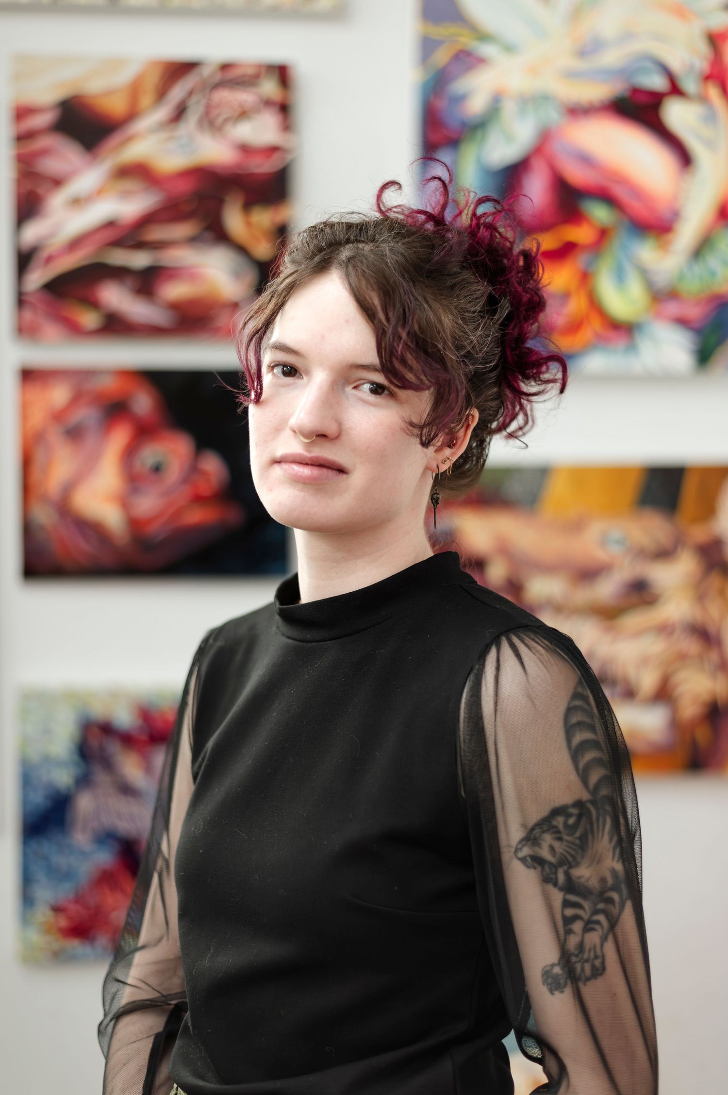 Person in black top with sheer sleeves and burgundy hair poses in front of vibrant abstract artwork.