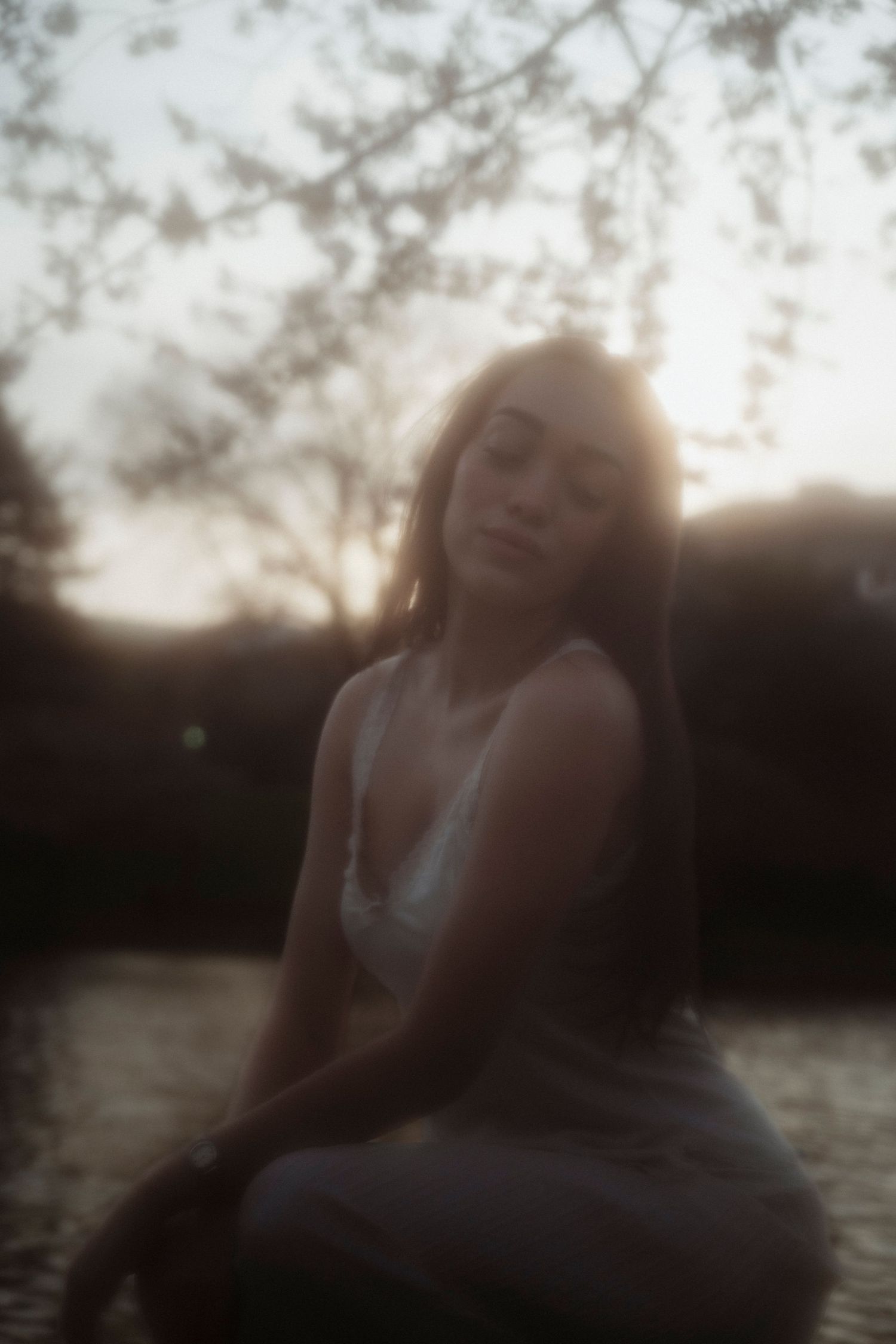 Young woman in white dress sitting outdoors at golden hour with soft bokeh sunset background.