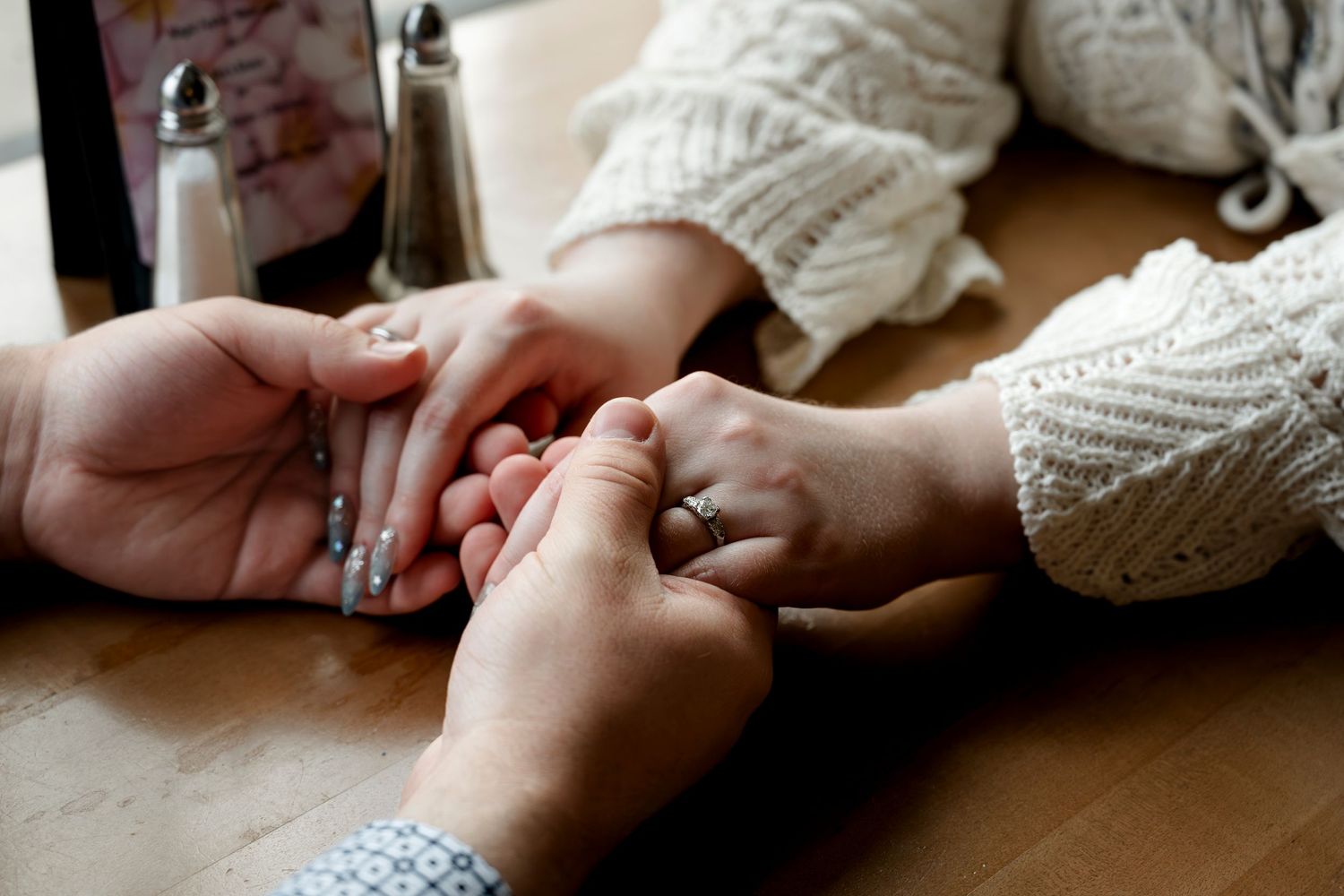 Two people holding hands tenderly across a wooden table, one wearing a knit sweater and a ring.