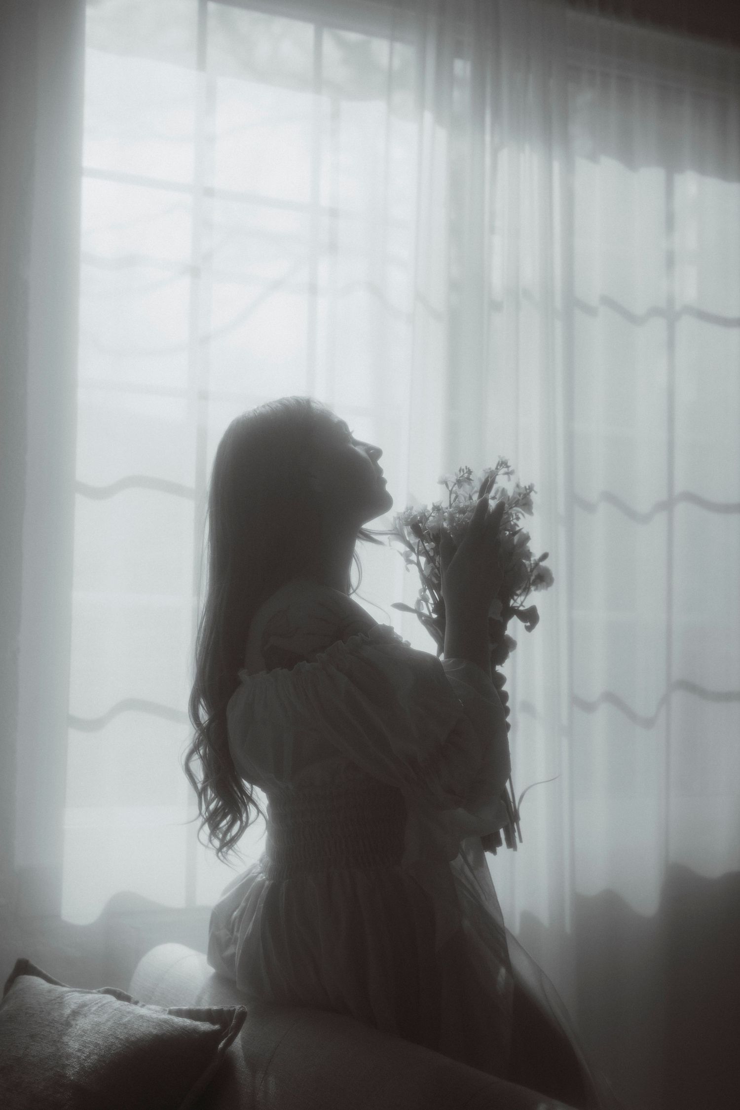 Silhouette of a woman in white dress holding wildflowers by sheer curtained window in moody black and white photo.