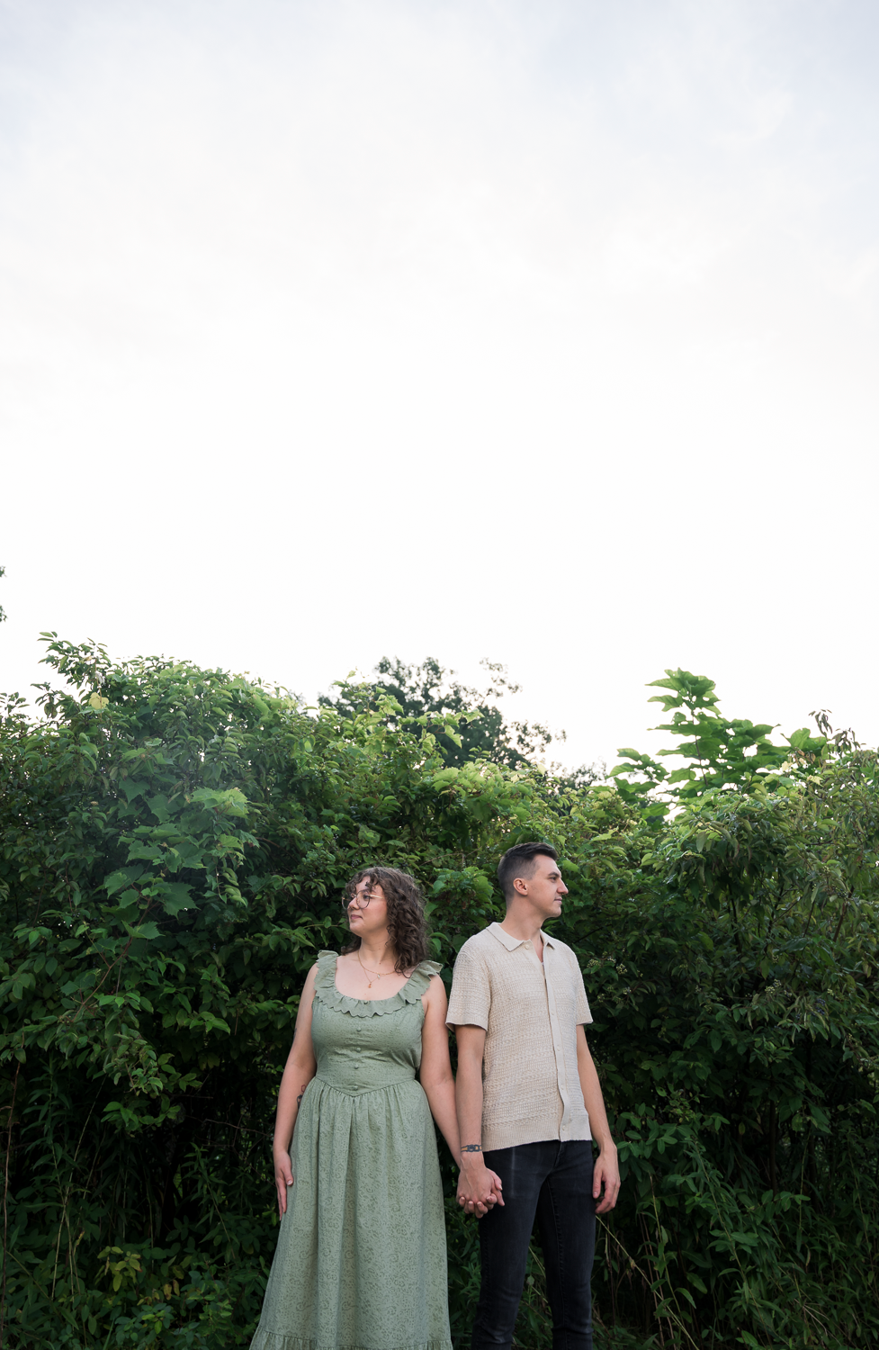 A couple holds hands while standing among tall green bushes, gazing upward in a serene outdoor setting.