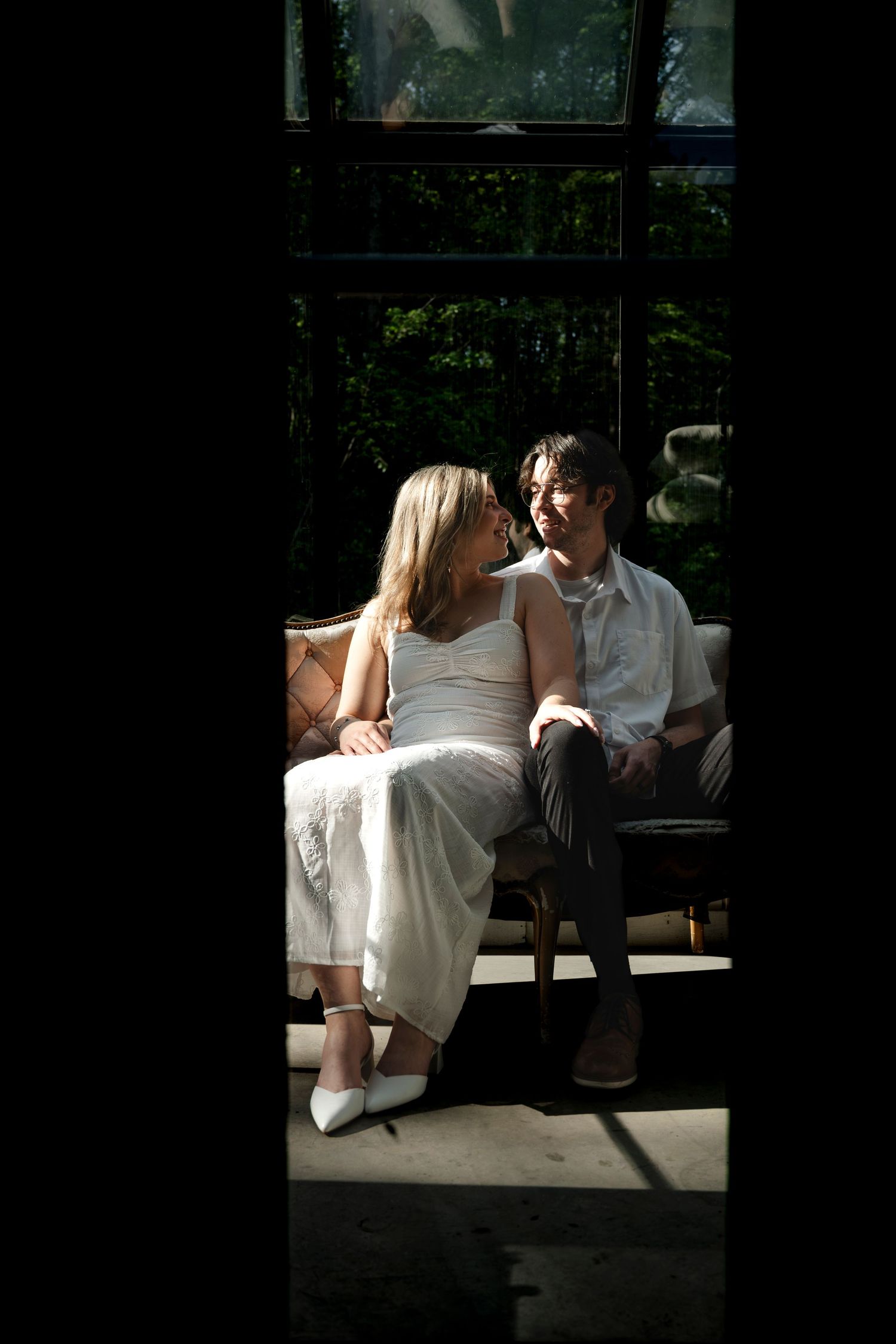 A couple sits close together on a sofa by a sunlit window, smiling at each other in elegant attire.
