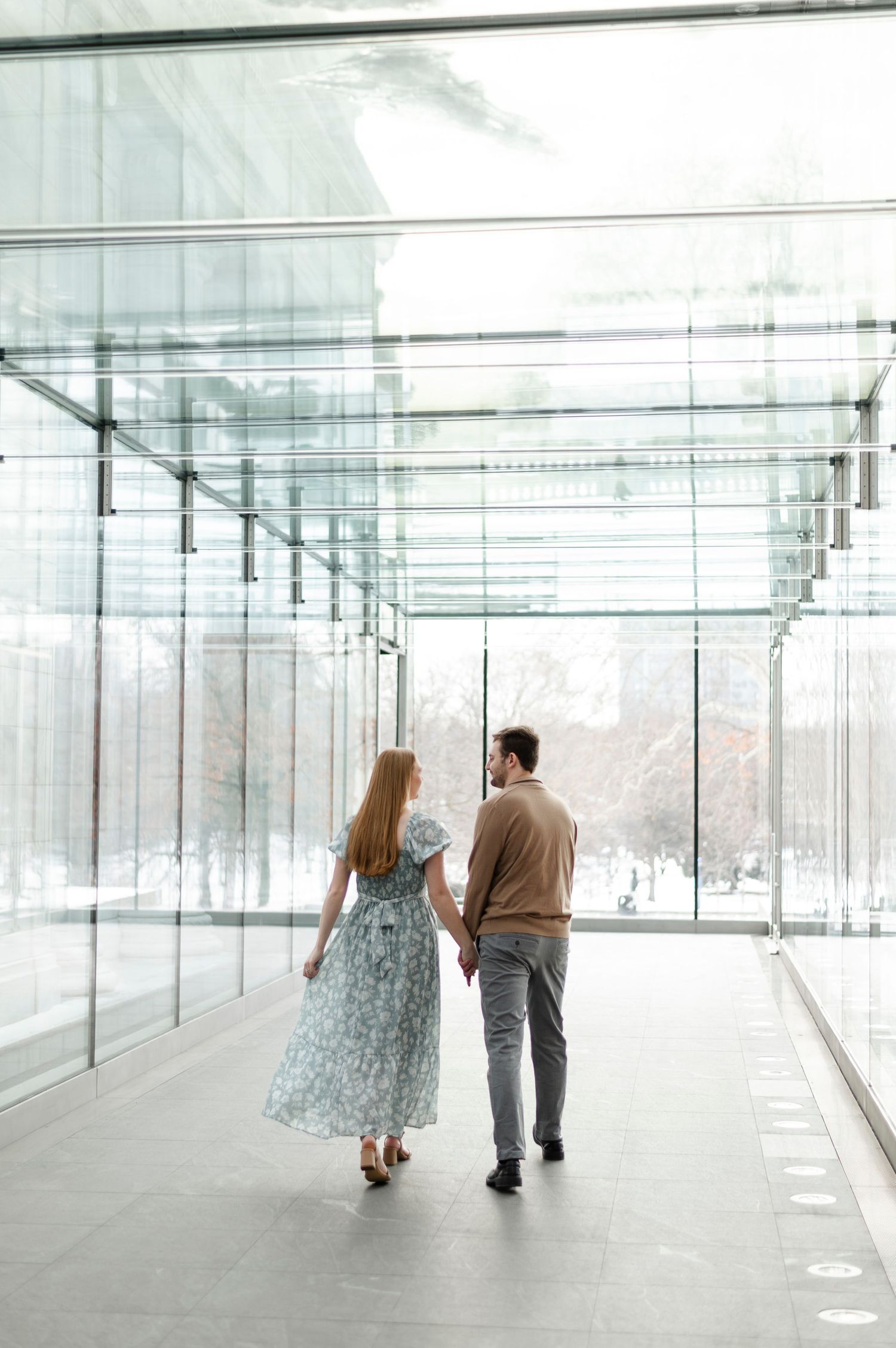 Couple walking hand in hand through modern glass corridor, woman in floral midi dress, man in tan sweater.