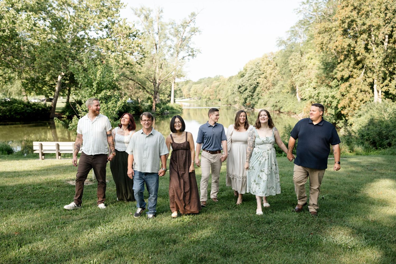 Group of eight people smiling and walking together on green grass near a serene pond surrounded by trees.