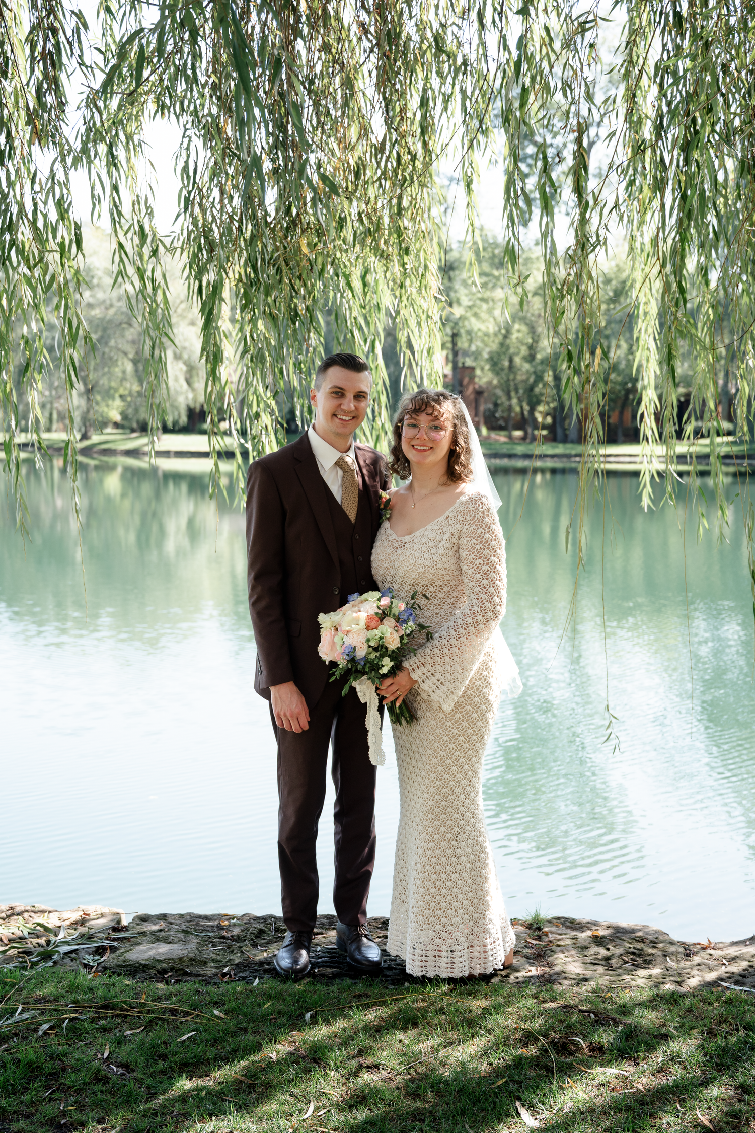 A couple poses for romantic wedding photos by a serene lake with hanging willow trees.