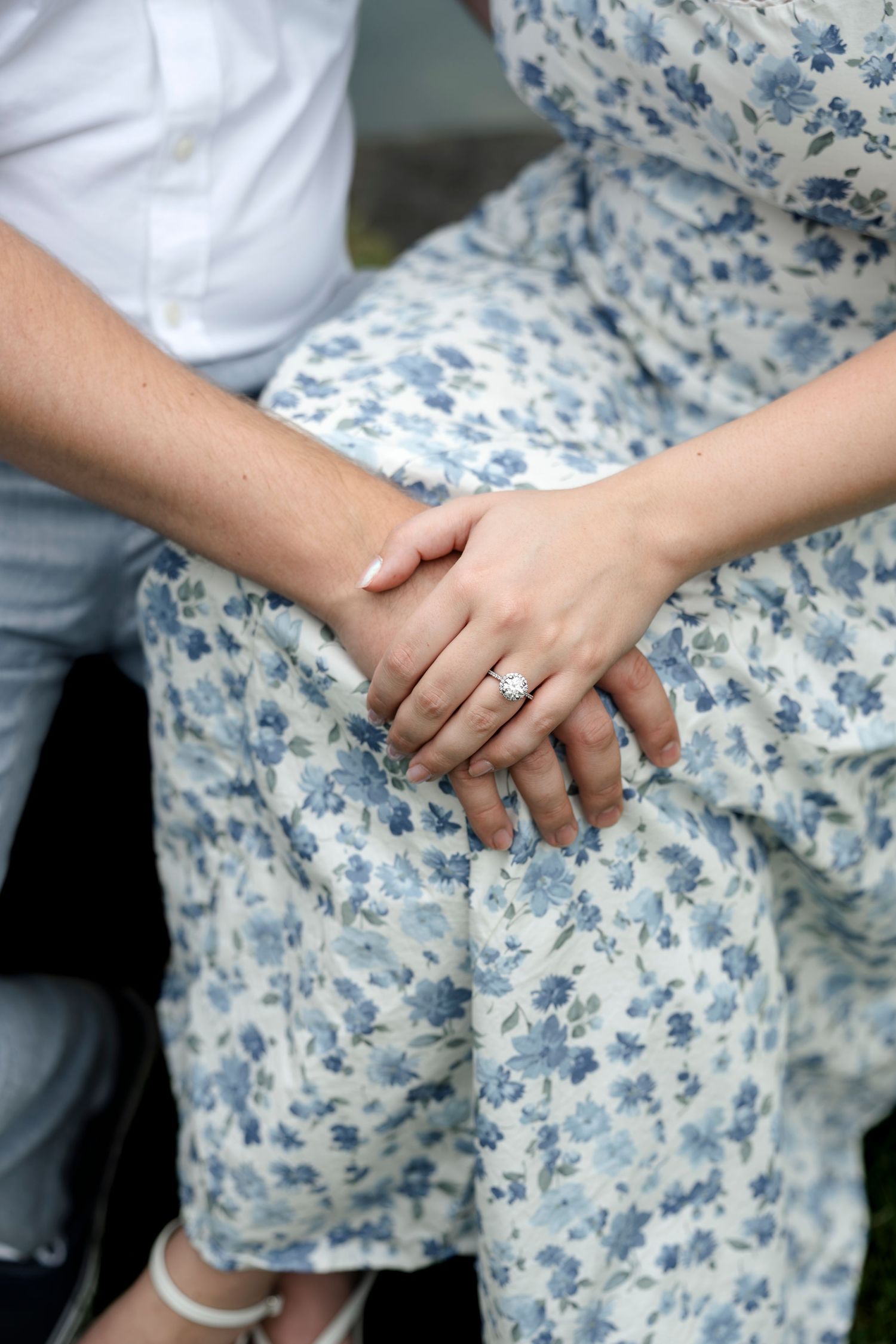 Close-up of couple holding hands, woman wearing diamond engagement ring, sitting together in blue floral dress.