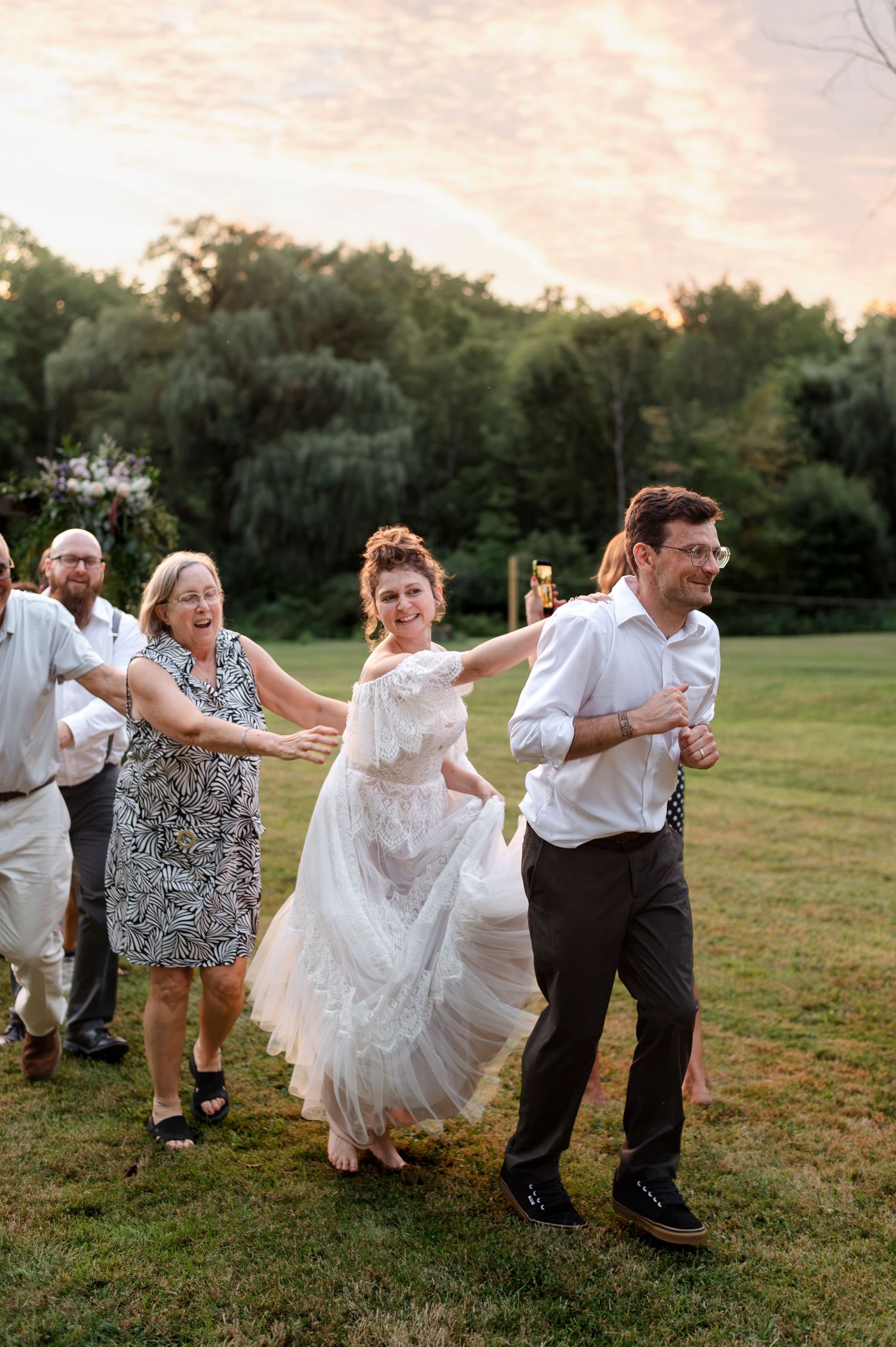 Bride in white gown dances outdoors with guests at sunset wedding reception on green lawn.