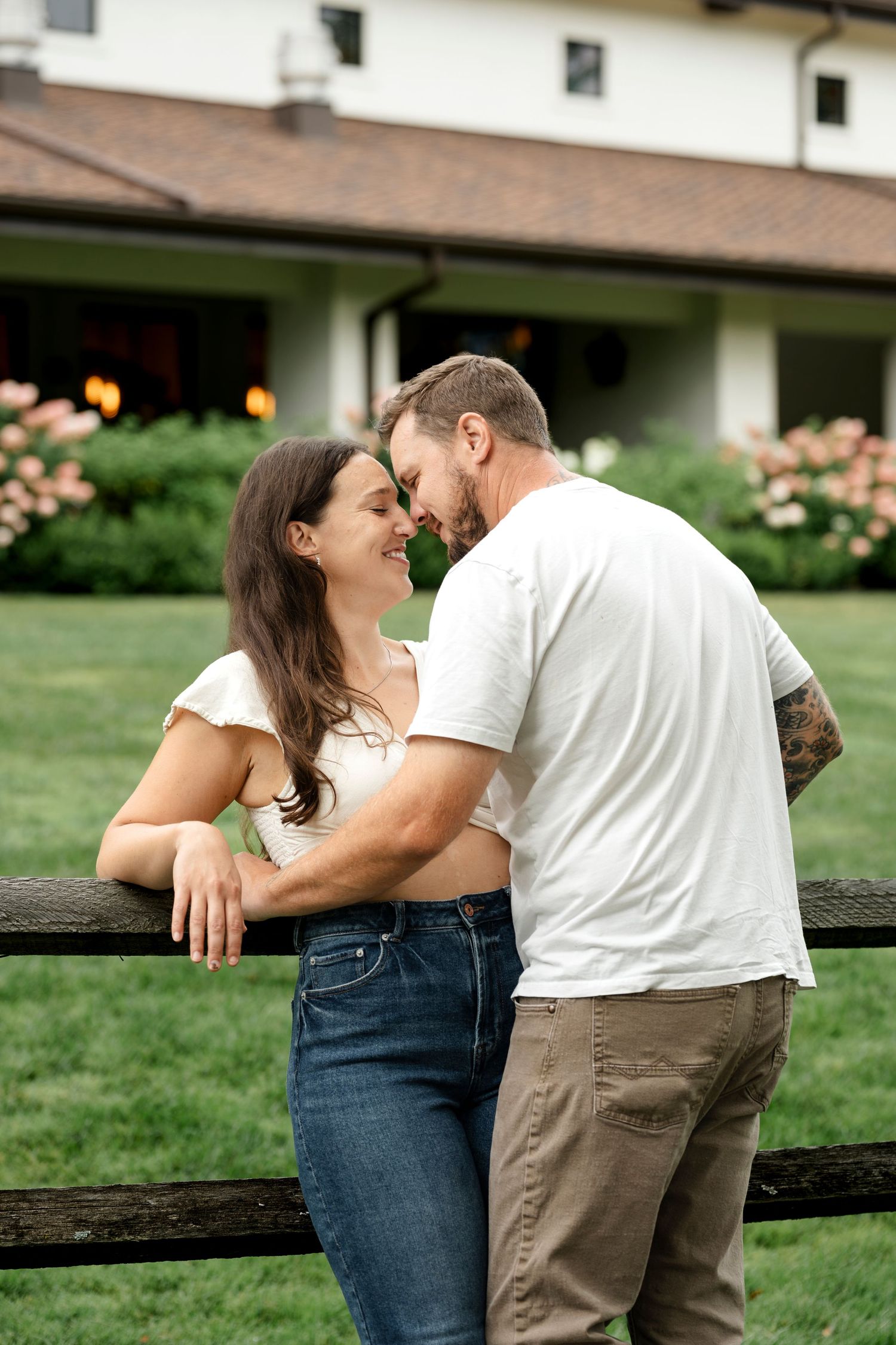 A couple shares a romantic moment leaning against a wooden fence with a green lawn and blooming flowers behind them.