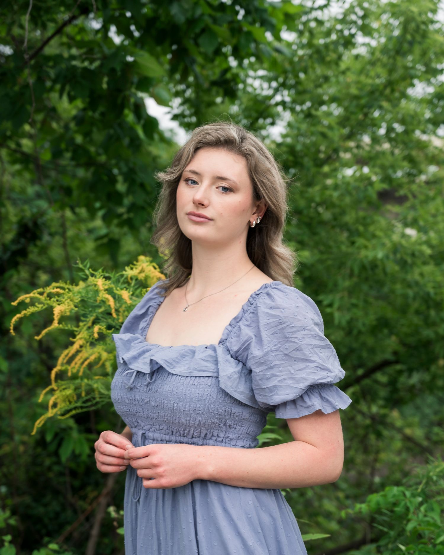 A person in a blue puff-sleeve dress poses outdoors against a green leafy background.