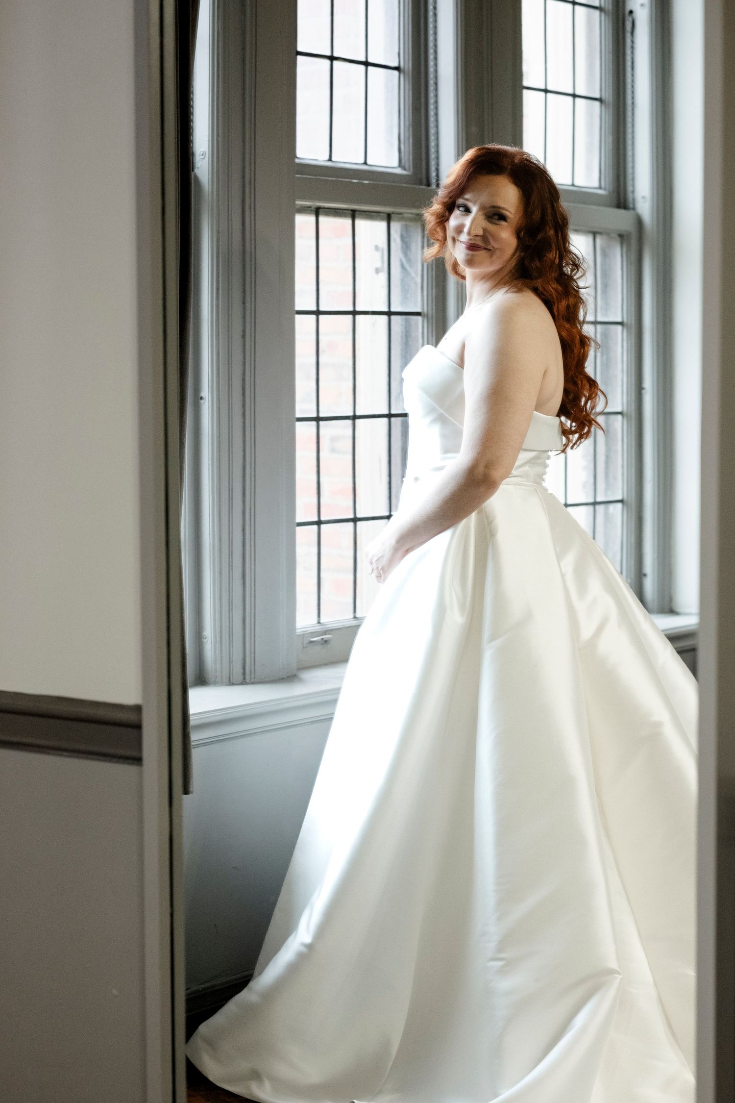 A bride in an elegant white wedding gown poses by a tall window with natural light streaming in.