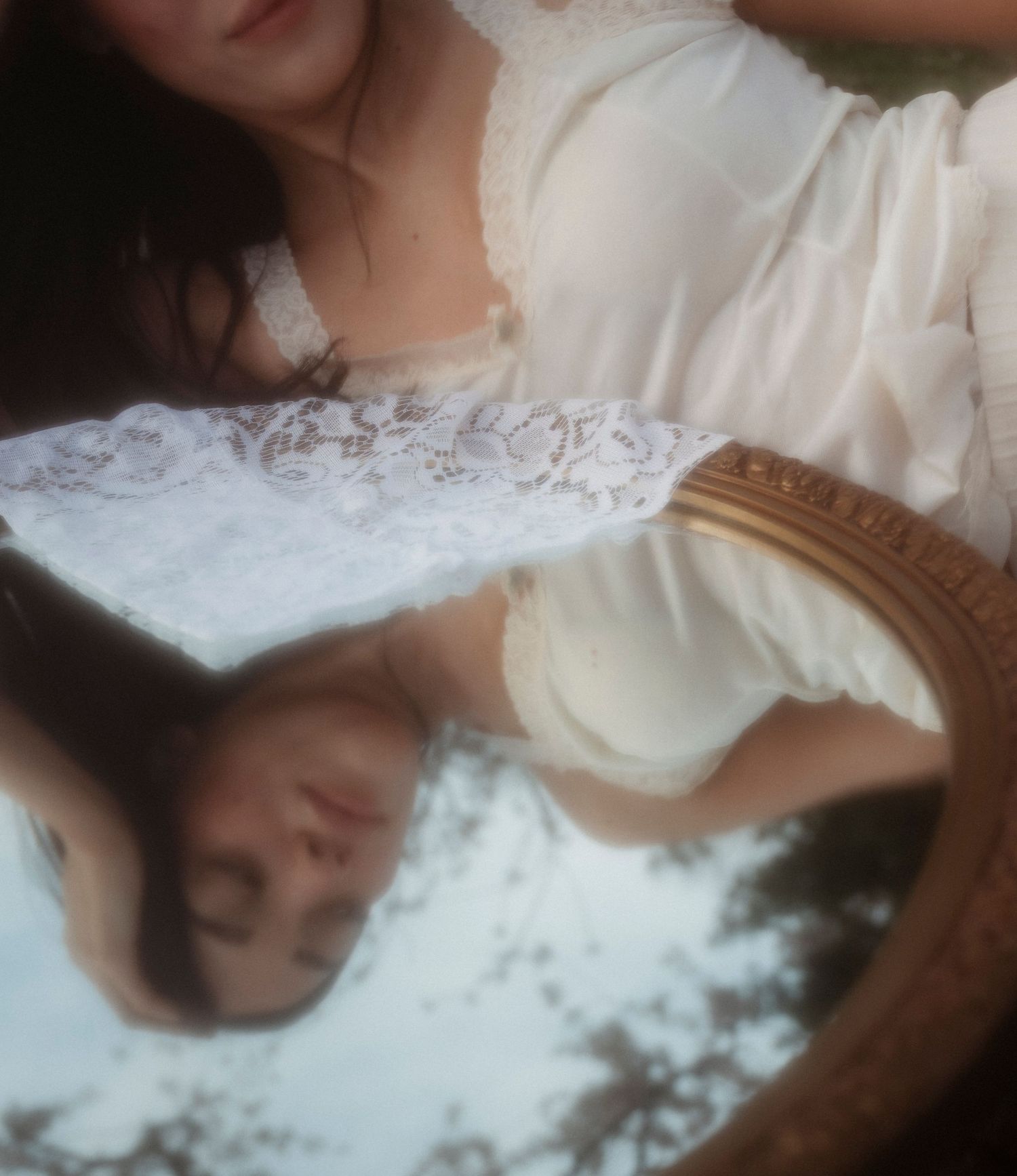 Young woman in white lace dress gazing into a decorative mirror reflecting her image outdoors.