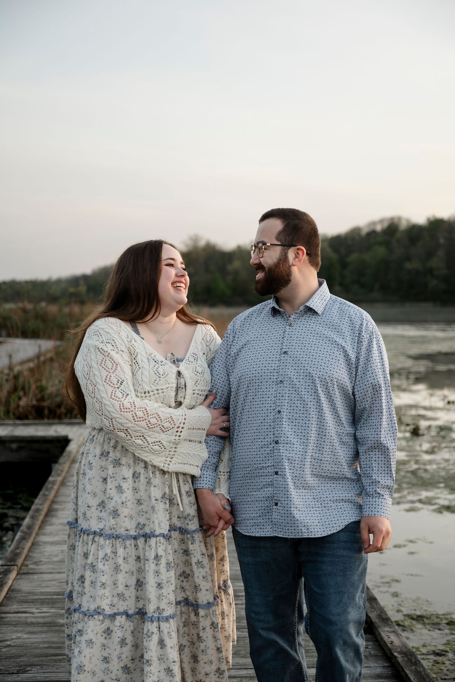 Smiling couple holding hands on a wooden dock with a serene lake and reeds in the background at dusk.