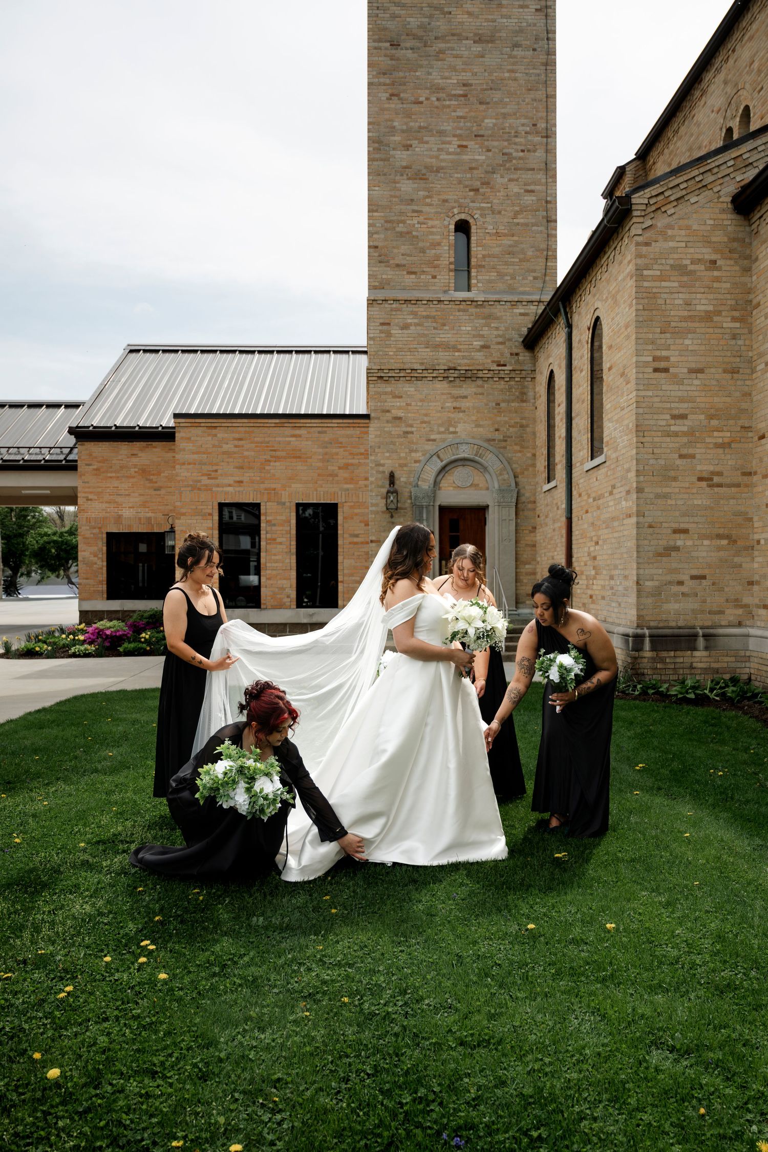 Bride in white gown with bridesmaids in black dresses outside a brick church, adjusting veil and holding bouquets.