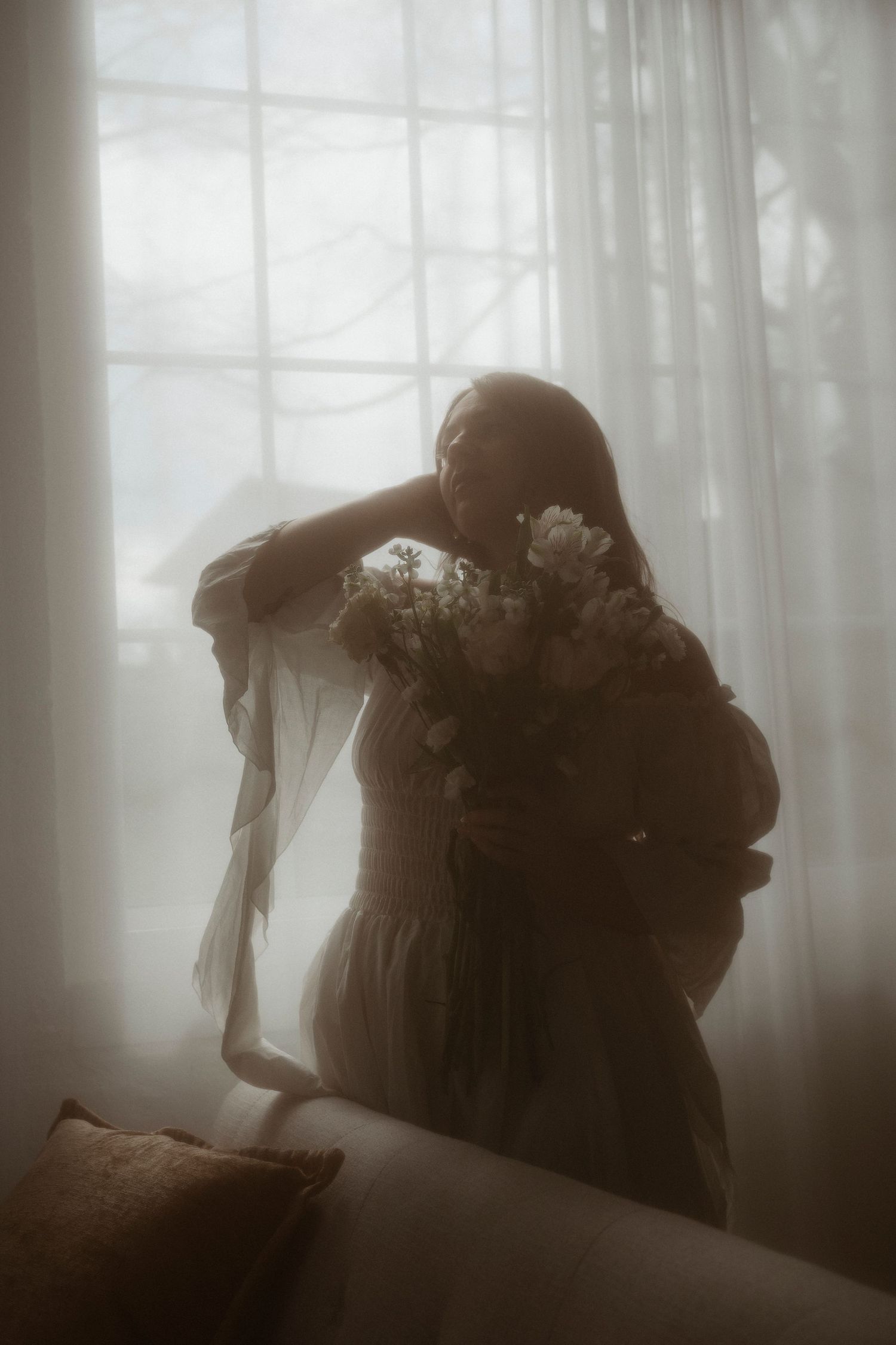Dreamy silhouette of a woman in a flowing dress holding a bouquet near sheer white curtains in soft light.