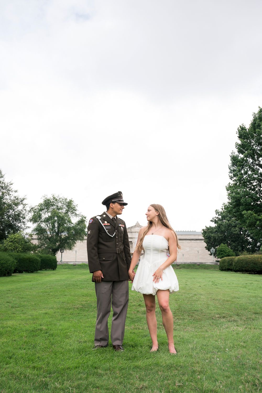 Military man in uniform holds hands with woman in white dress on green lawn near historic building.