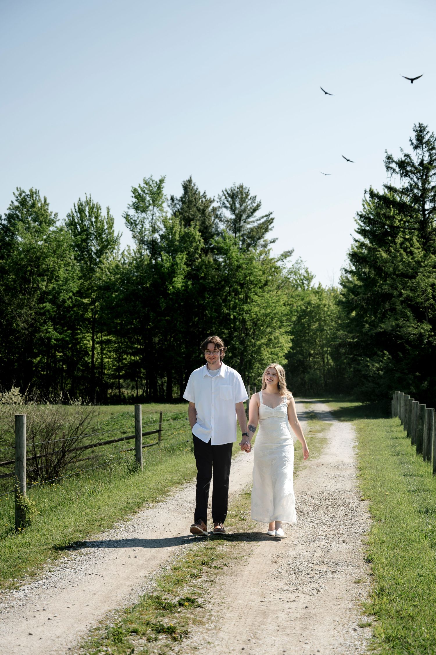 Bride and groom holding hands walking down sunny gravel path lined with green trees on their wedding day.