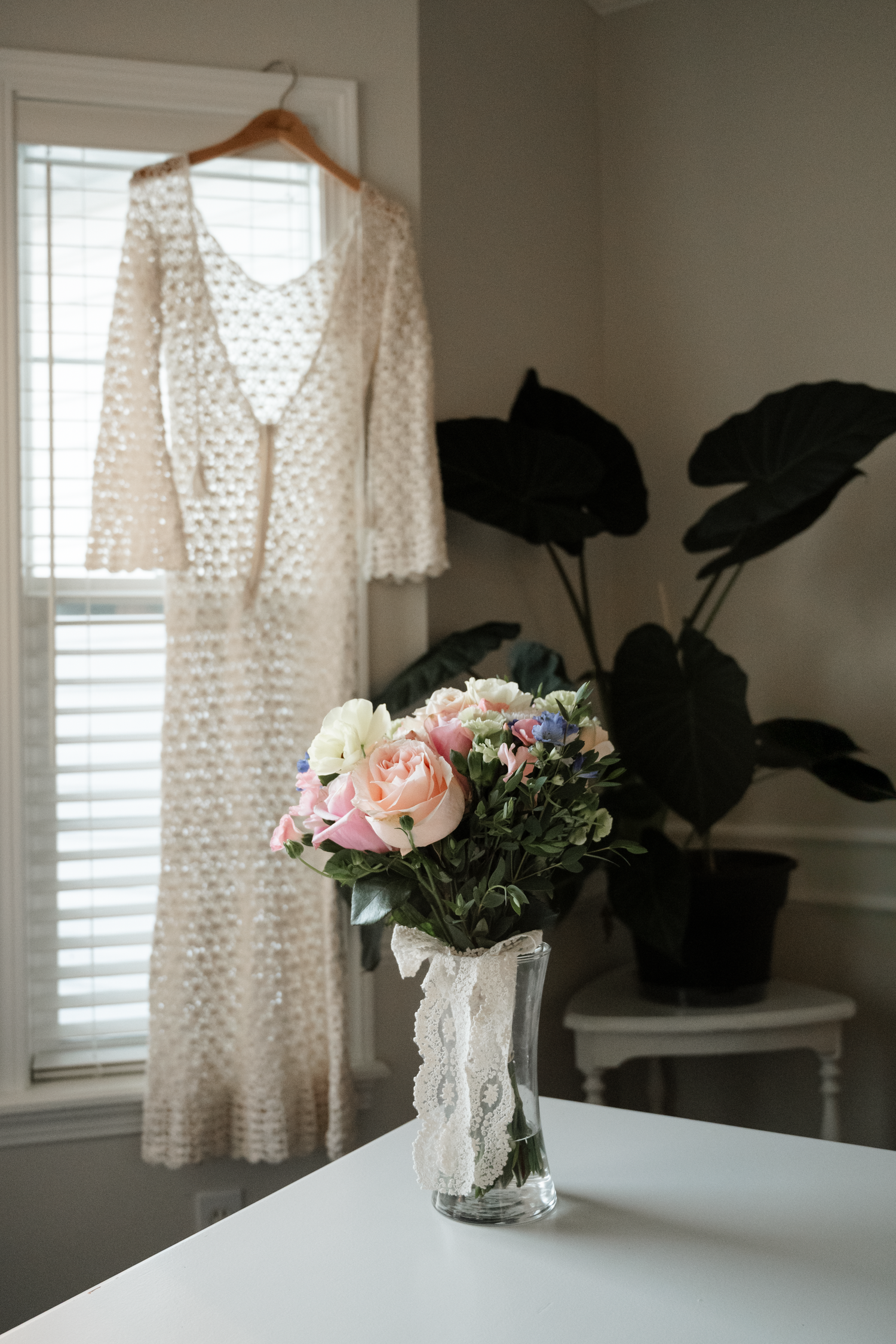 A delicate white lace dress hangs by a window with soft pink roses in a vase on a nearby table.