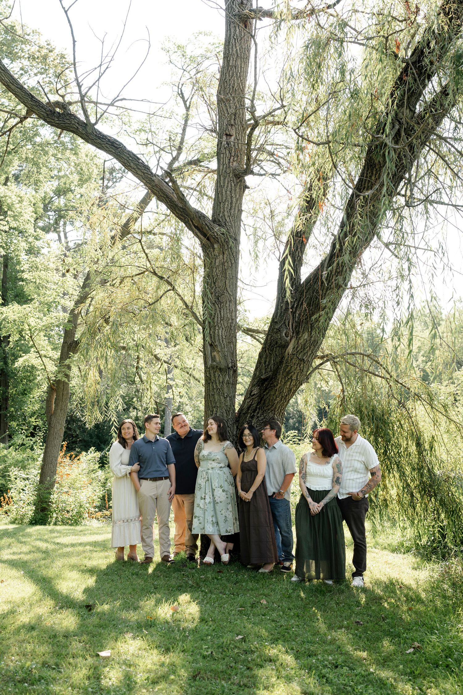 A family of eight poses together under a large willow tree in a lush green garden on a sunny day.