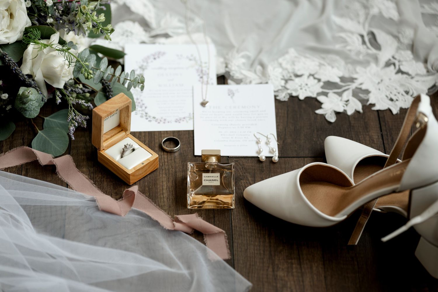 Elegant bridal flat lay with white heels, perfume, wedding rings, floral bouquet, and invitation on wood.